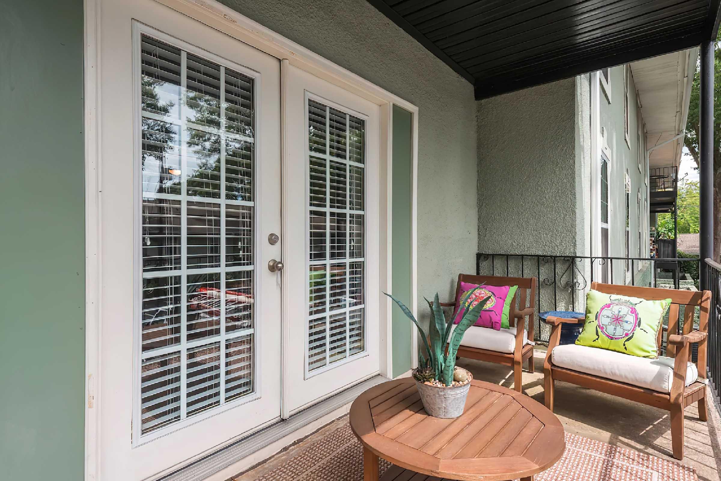 A cozy balcony featuring white French doors with glass panes, flanked by two comfortable chairs adorned with colorful cushions. A round wooden coffee table sits in the center, topped with a potted plant. The balcony is surrounded by a railing and has a pleasant, inviting atmosphere.