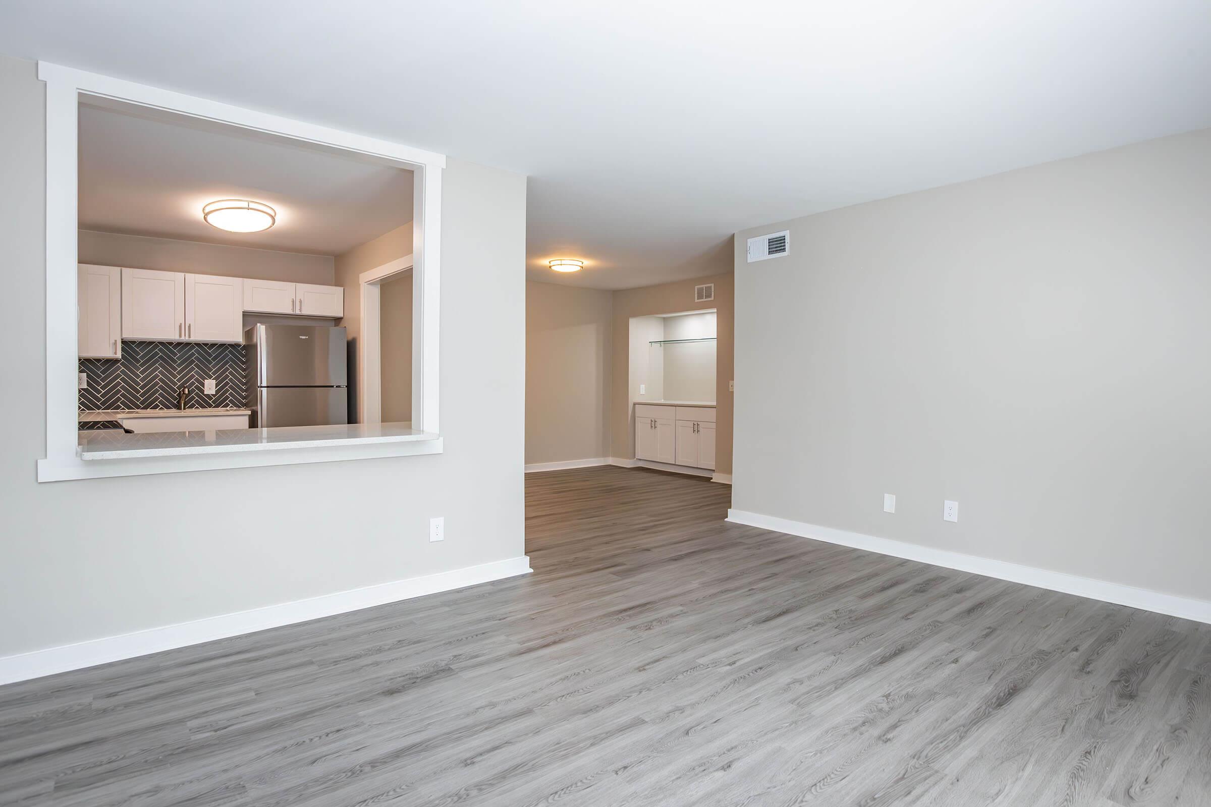 A spacious, modern apartment interior featuring light gray walls and wood-like flooring. To the left, a kitchen area is visible through an opening with white cabinetry and a stainless steel fridge. The right side shows an empty living area with ample natural light.