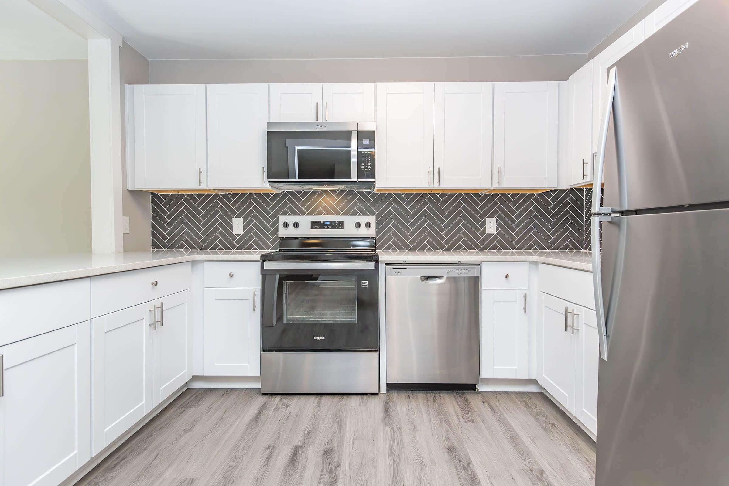 Modern kitchen featuring white cabinetry, a stainless steel refrigerator, oven, and dishwasher. The backsplash is designed with herringbone-patterned tiles, and the countertops are light-colored. The flooring is laminate in a wood-like finish, creating a clean and contemporary look.