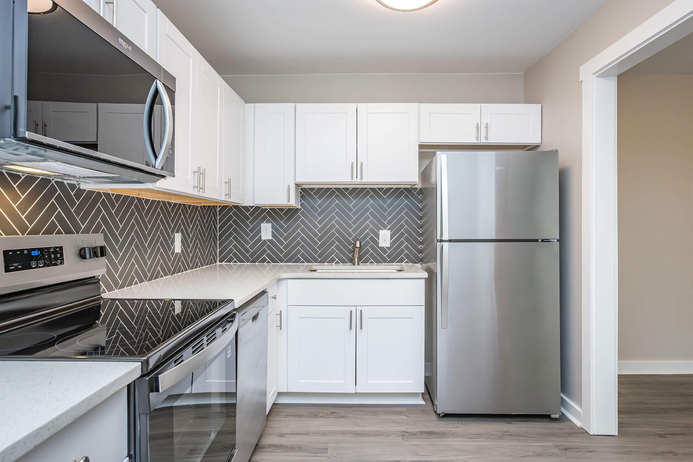 Modern kitchen with white cabinetry, stainless steel appliances including a refrigerator and microwave, and a sleek countertop. The backsplash features a herringbone tile pattern. The space is well-lit and designed with a contemporary aesthetic, featuring a neutral color palette and laminate flooring.