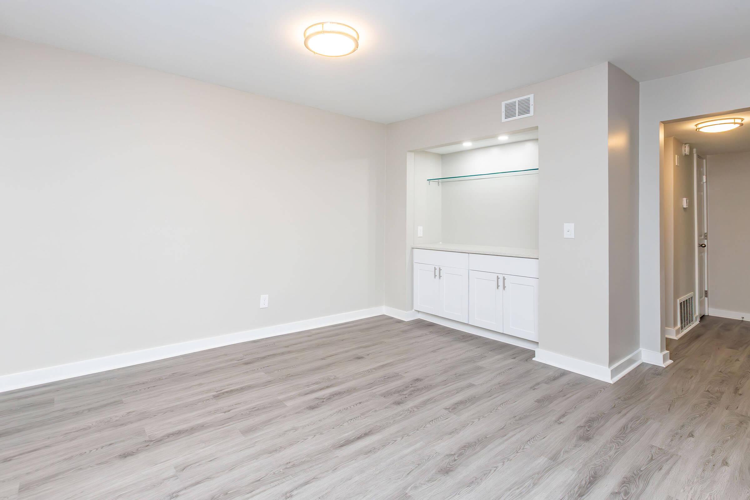 A vacant, well-lit interior space featuring light gray walls and a wood-like floor. There is a small built-in shelving unit with cabinets along one wall, illuminated with soft lighting. The layout is open and uncluttered, with a doorway visible on the right leading to another room.