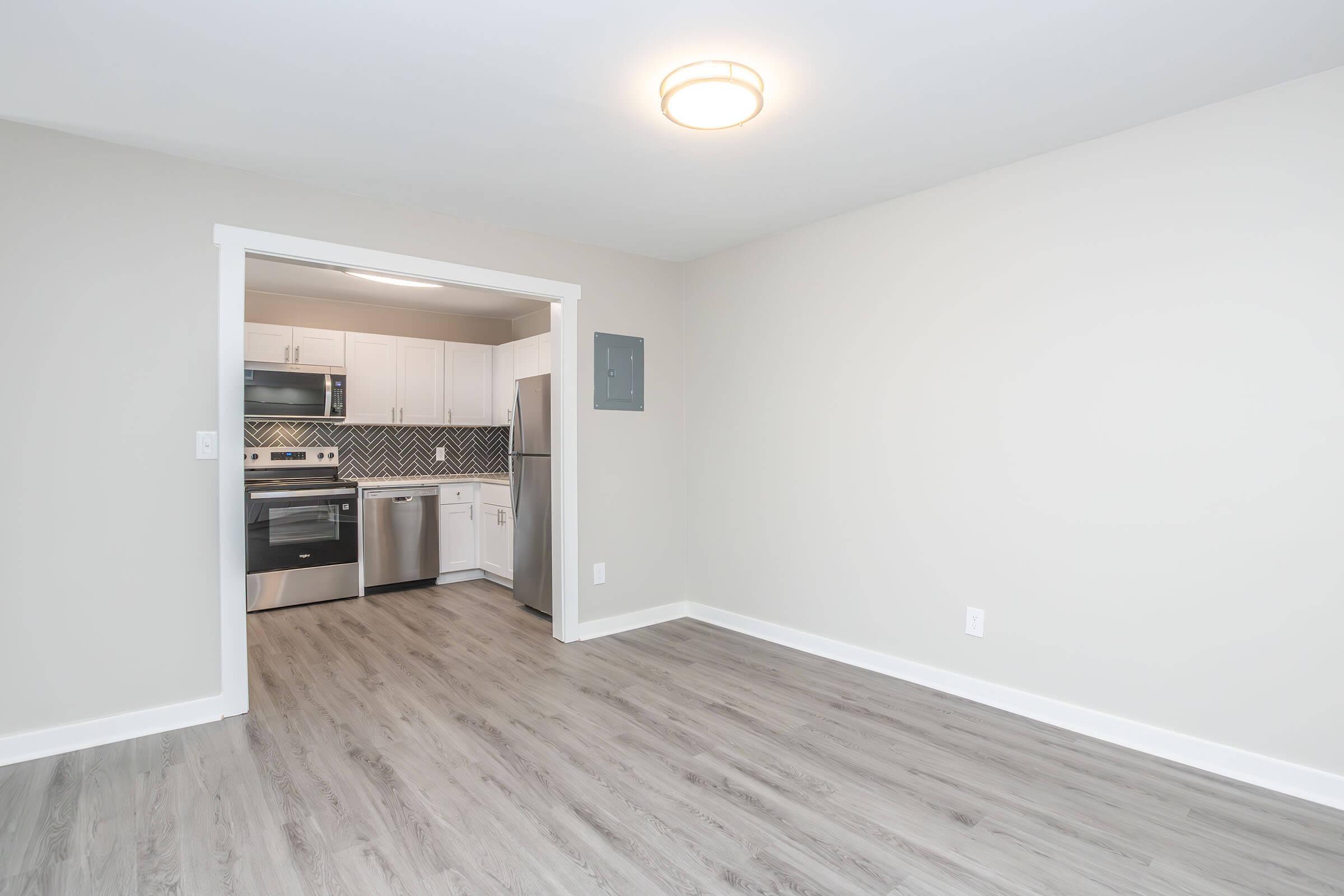 Spacious, modern kitchen seen from a living area. Features white cabinetry, stainless steel appliances, and a decorative backsplash. The floor is light gray, creating a cohesive look with the neutral wall color. Ample natural light from the ceiling fixture enhances the open layout.