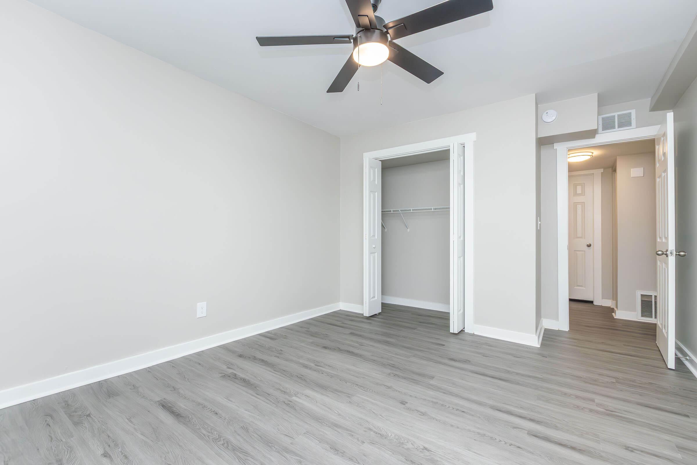 Empty room with light gray walls and modern laminate flooring. A ceiling fan is mounted on the ceiling, and a white closet with double doors is visible on the left. There is another door on the right leading to a hallway, and the overall space is well-lit with natural light.