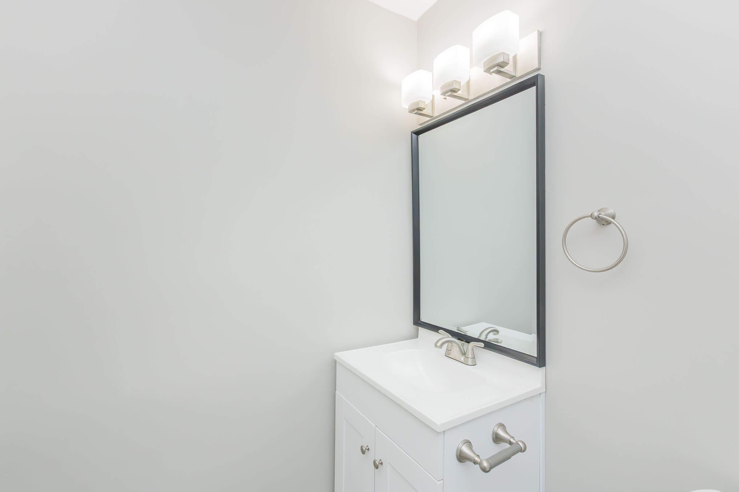 A modern bathroom featuring a white vanity with a sink, a large rectangular mirror framed in black, and three light fixtures above. A towel ring is mounted on the wall, and the wall is painted in a light neutral color. The space is clean and minimalistic, emphasizing simplicity and elegance.