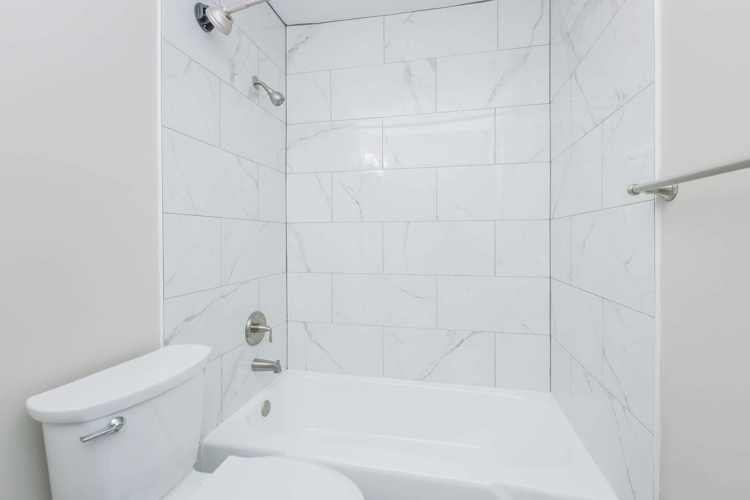 A modern bathroom featuring a white bathtub, tiled walls with a marble design, a stainless steel showerhead, and a toilet. The space is bright and clean, with neutral colors and minimal fixtures.