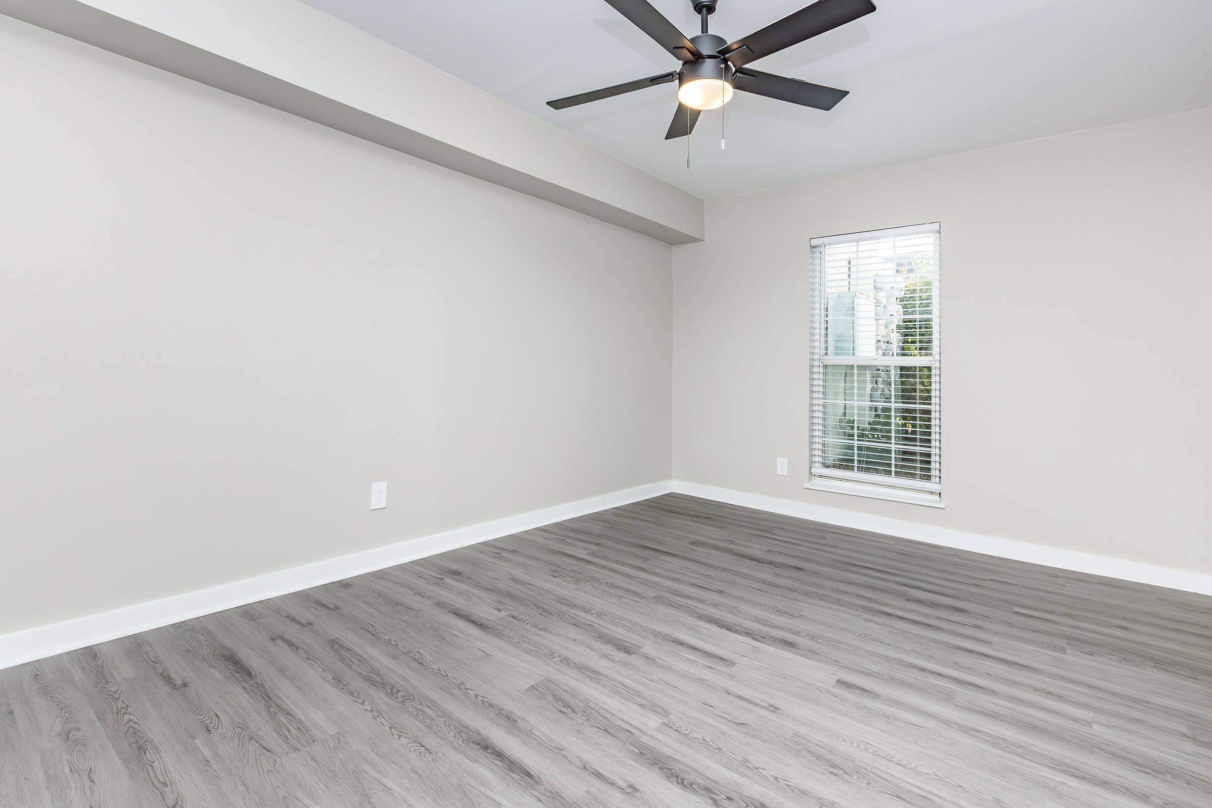 A spacious, empty room with light gray walls and a light wood-style floor. There is a ceiling fan with black blades, and a window with white blinds letting in natural light. The room has a clean, modern feel, and is ready for decoration or furniture placement.