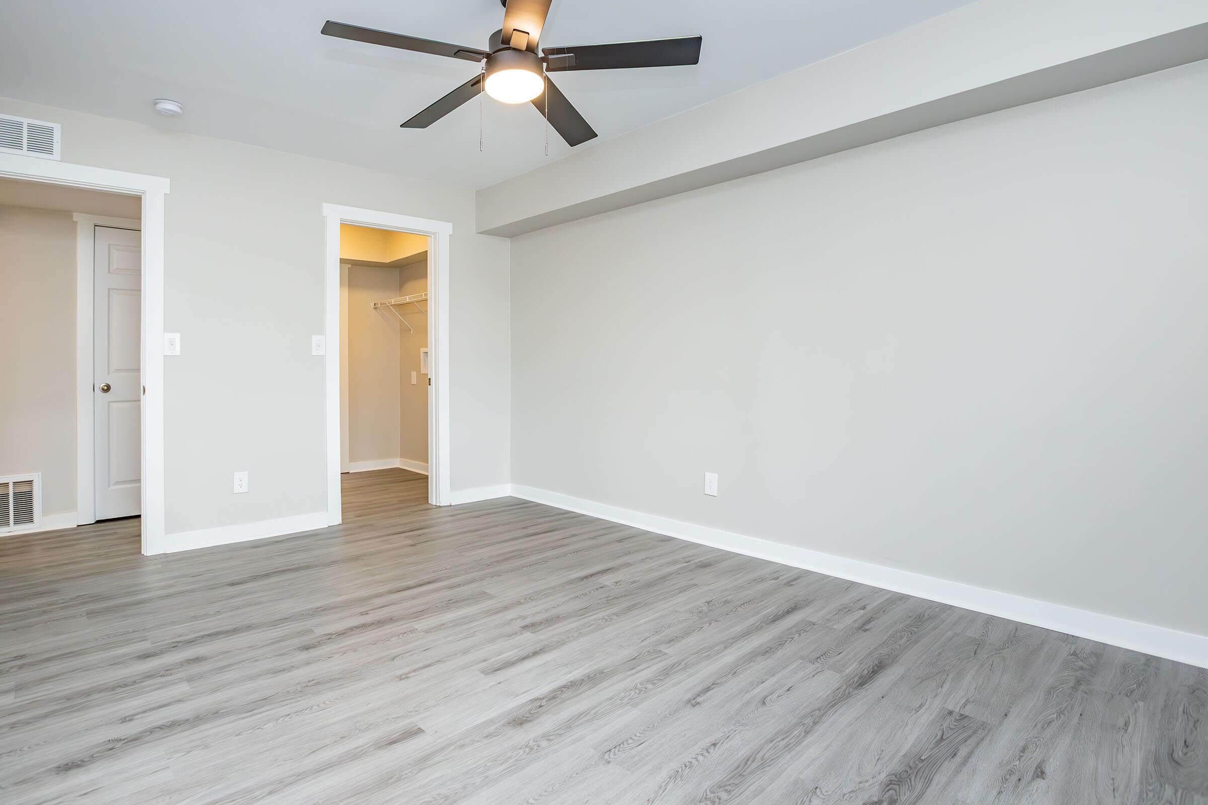 A spacious, empty room with light gray walls and a wood-like floor. A ceiling fan is mounted in the middle, and there's a doorway leading to a small closet. The room has natural lighting, creating a bright and airy atmosphere.