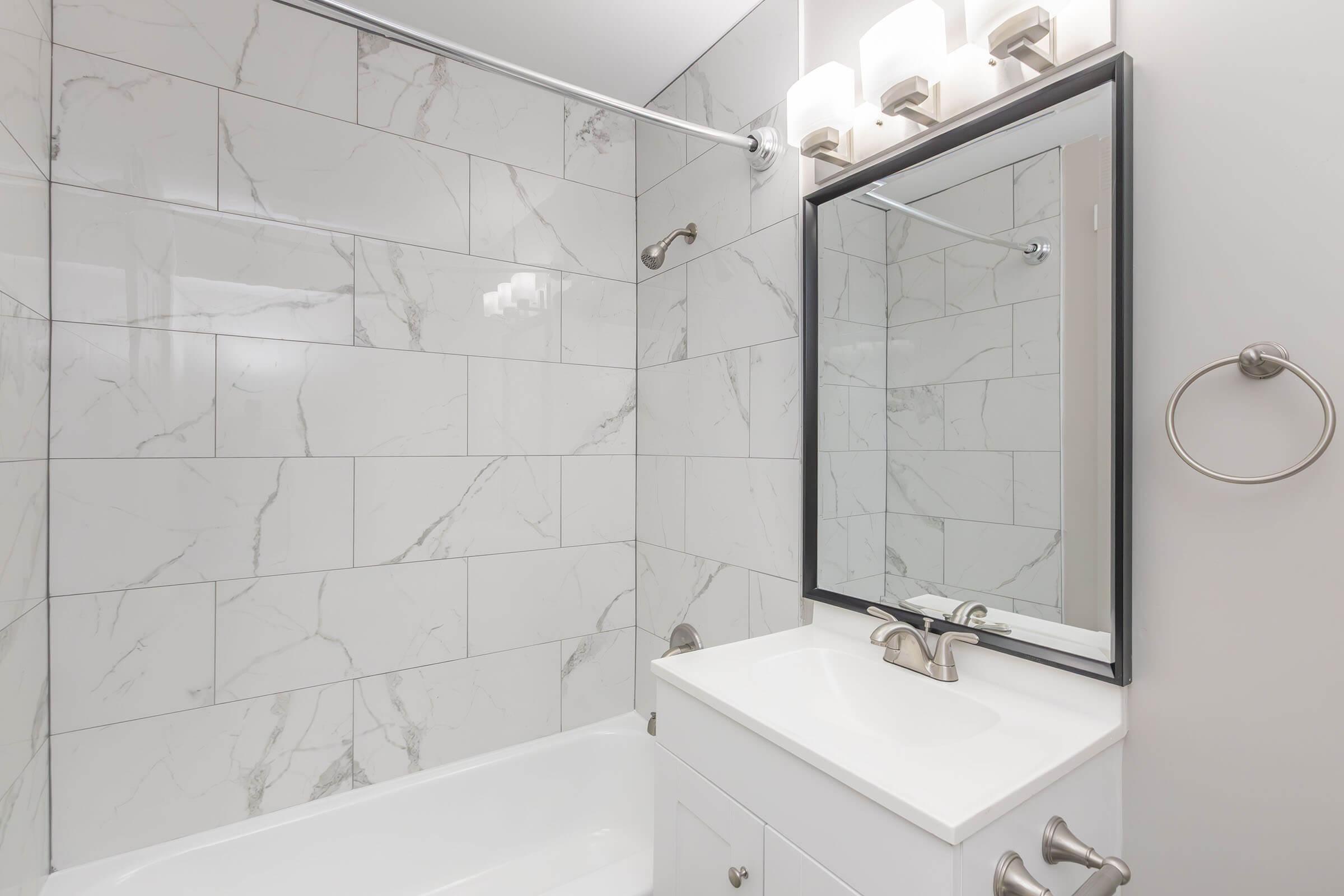 A modern bathroom featuring white marble-patterned tiles, a bathtub, a white vanity with a sink, and a large black-framed mirror above the sink. The lighting consists of four sconces mounted above the mirror. A chrome towel ring is visible on the wall next to the bathtub.