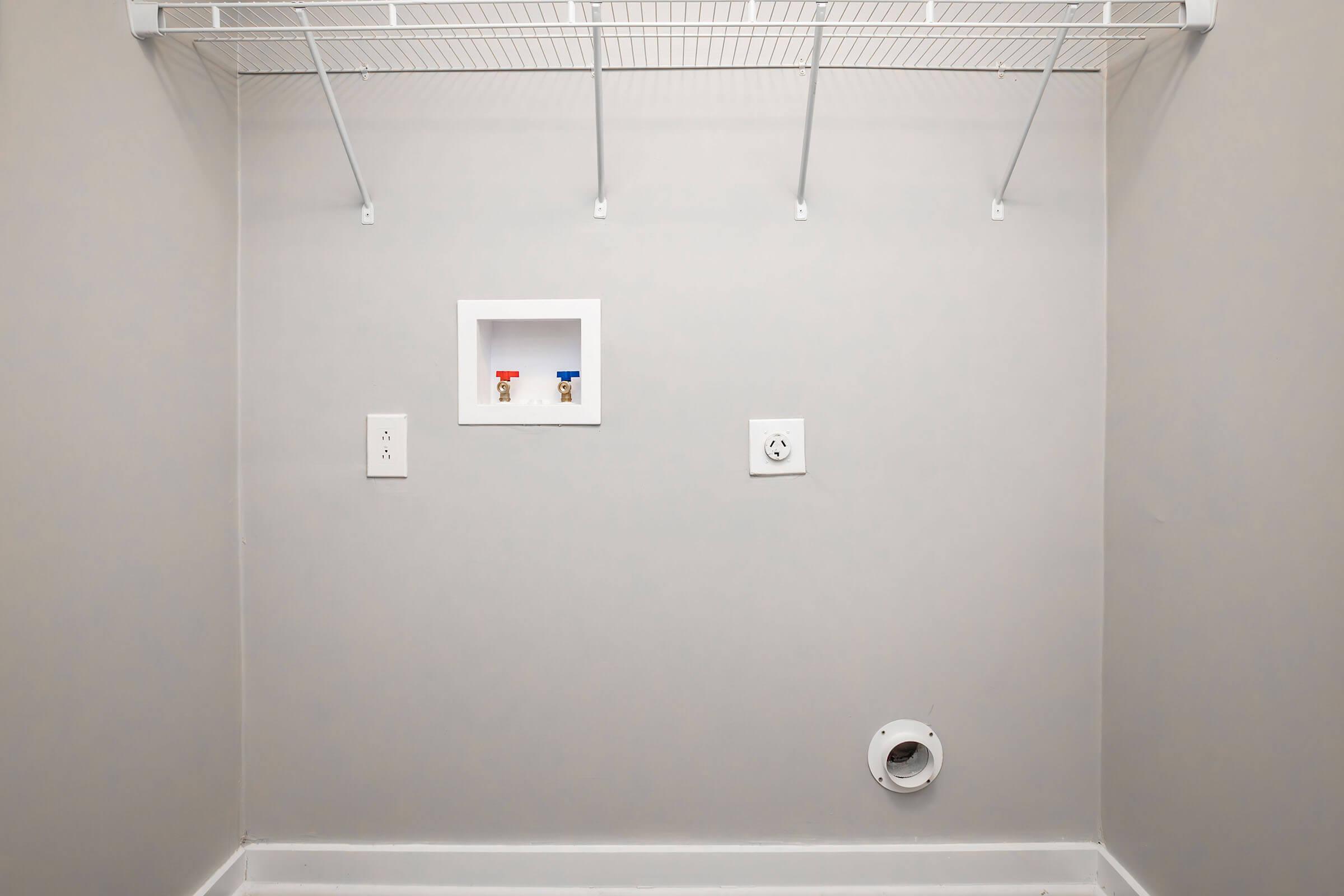 Empty laundry room with gray walls and a white floor. There are five wire shelves mounted on the wall, and a small white inset with two valves. Electrical outlets and a vent hole are visible on the wall. The space is well-lit and uncluttered, creating a minimalistic appearance.