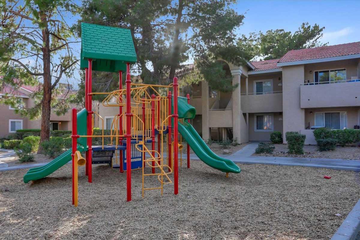 A colorful playground with a climbing structure, slides, and safety features, surrounded by tan sand and bordered by grass. In the background, there are residential buildings with balconies and greenery. The scene is bright and inviting, suitable for children to play.