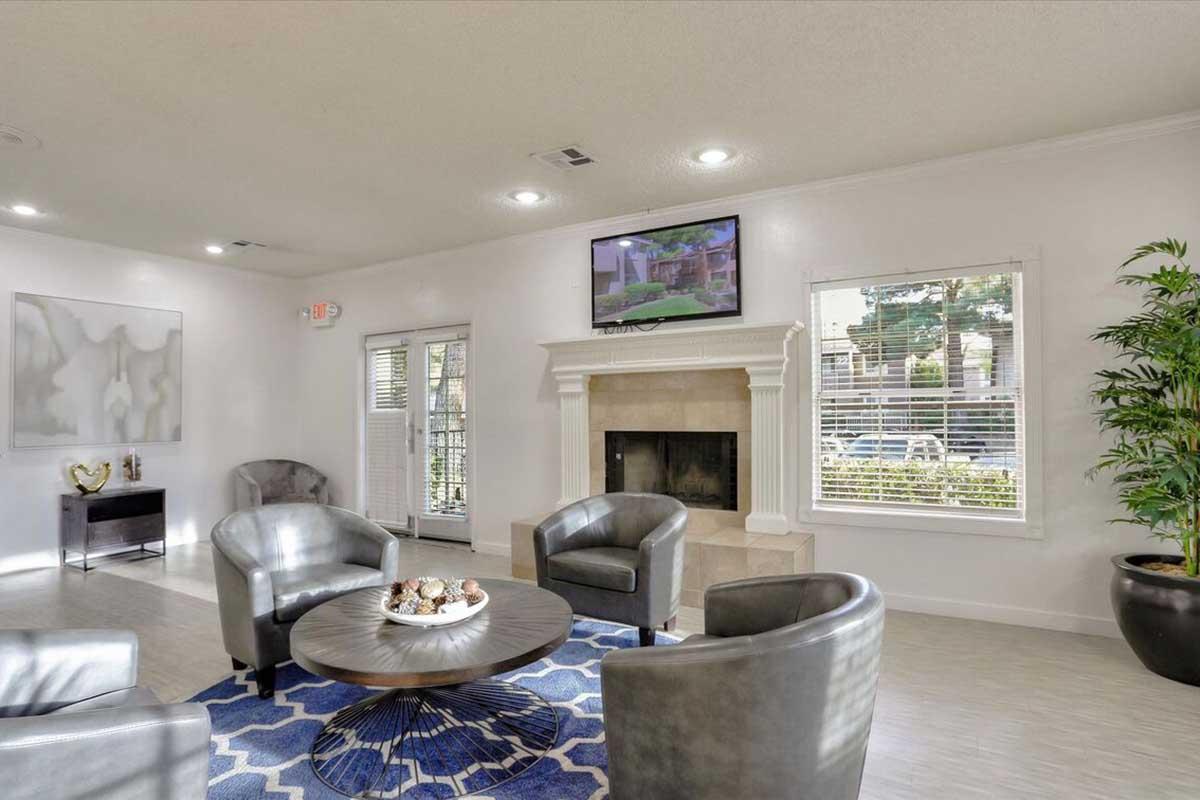 A modern living room with a cozy seating area featuring four gray chairs arranged around a circular table. A stone fireplace is in the background beneath a flat-screen TV. There are large windows with blinds, letting in natural light, and a decorative plant adds a touch of greenery to the space.