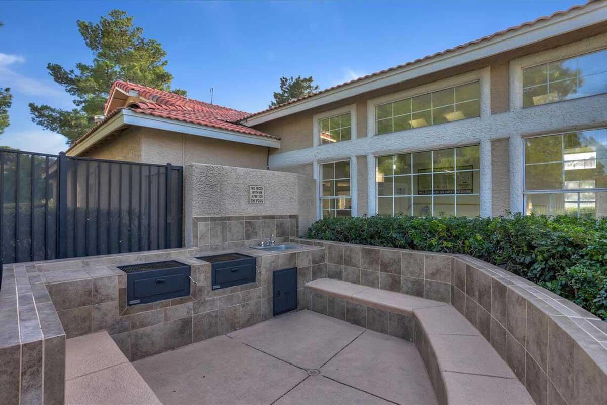 Outdoor communal space featuring a paved area with built-in seating and two BBQ grills, surrounded by greenery. The background includes a light-colored building with large windows and a red-tiled roof. A black gate encloses the area, adding security and privacy.