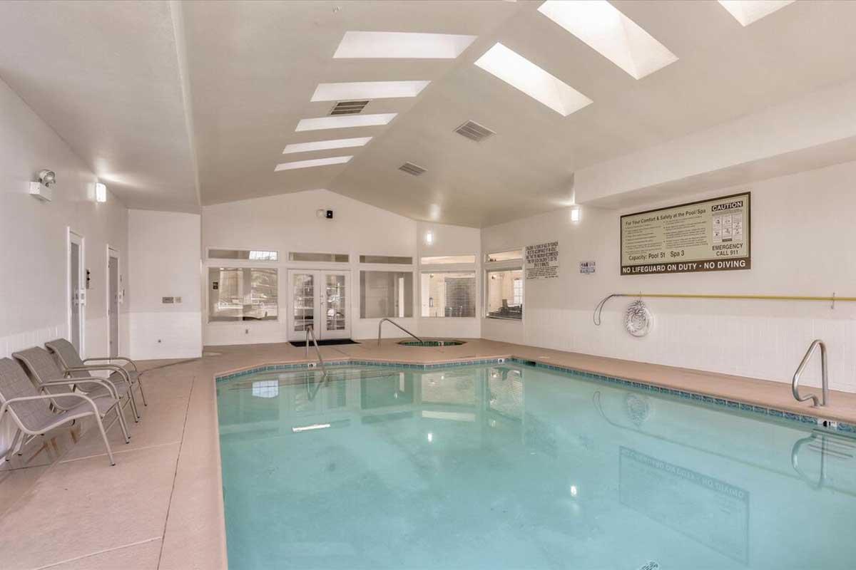 Indoor swimming pool area with a clear blue water, surrounded by white walls and large windows allowing natural light. There are a few lounge chairs on one side, and safety signs posted on the wall. The ceiling features skylights, enhancing the bright and airy atmosphere.