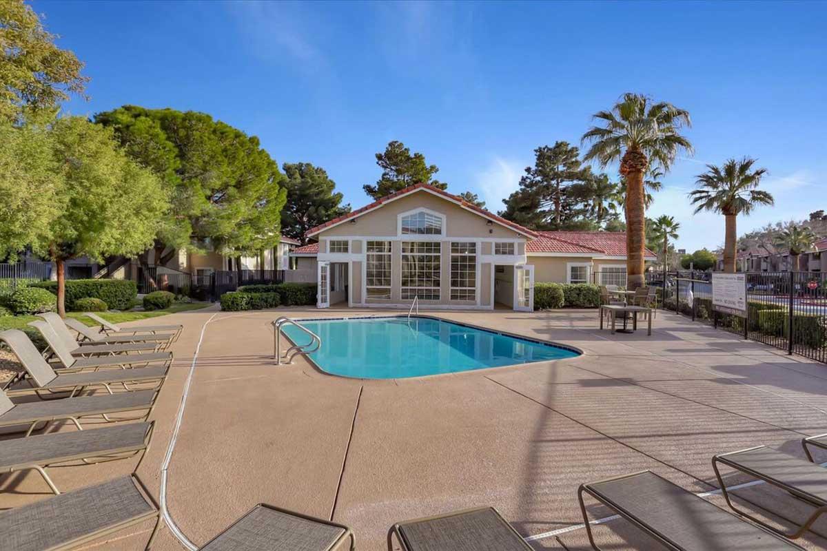 A spacious outdoor pool area featuring lounge chairs, surrounded by lush greenery and palm trees. The pool is located near a modern building with large windows, providing a welcoming atmosphere for relaxation and leisure activities.