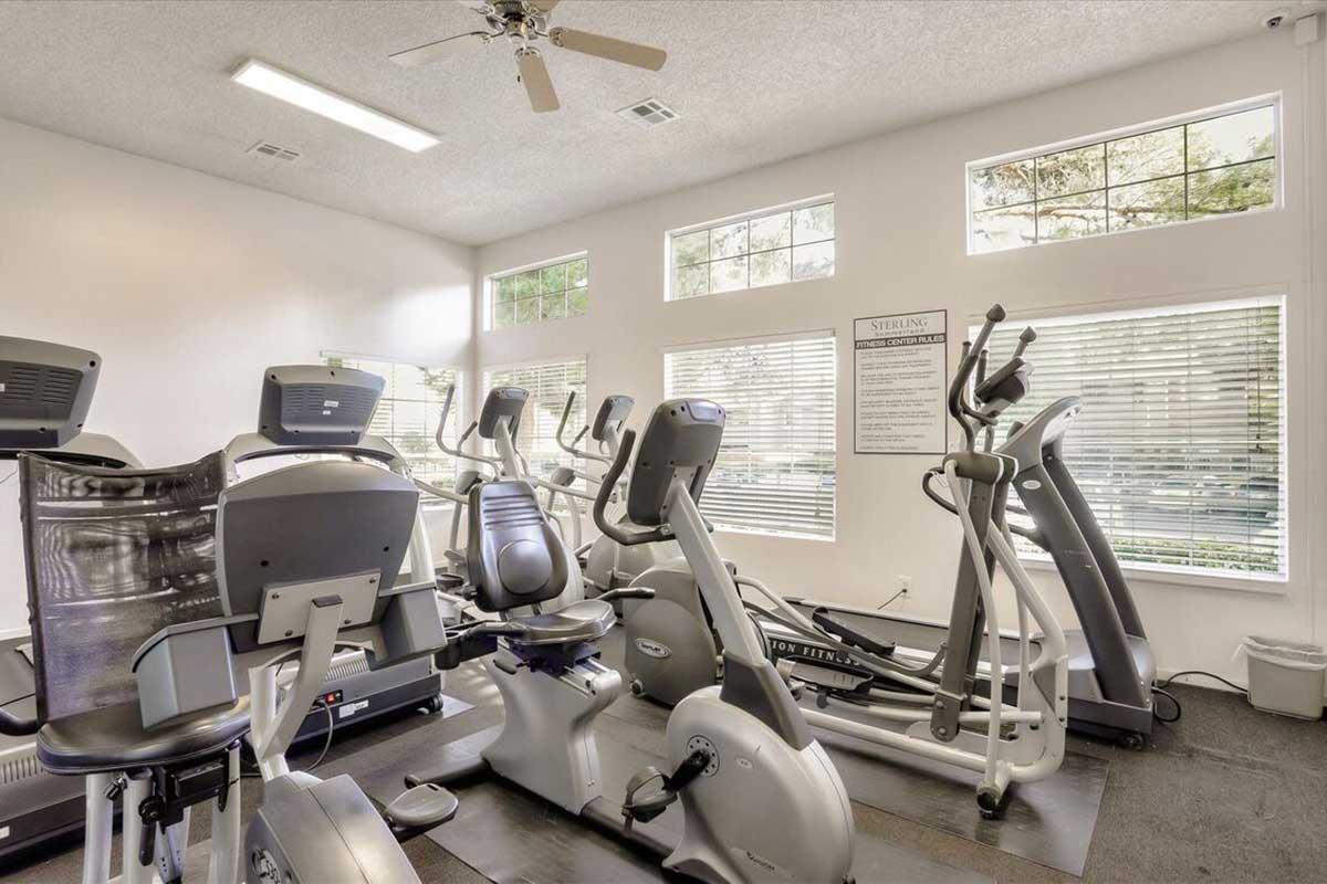 A bright gym interior featuring various exercise machines, including treadmills, elliptical trainers, and stationary bikes. Large windows allow natural light to fill the space, and a fan is visible on the ceiling. Wall-mounted information signage is also present.