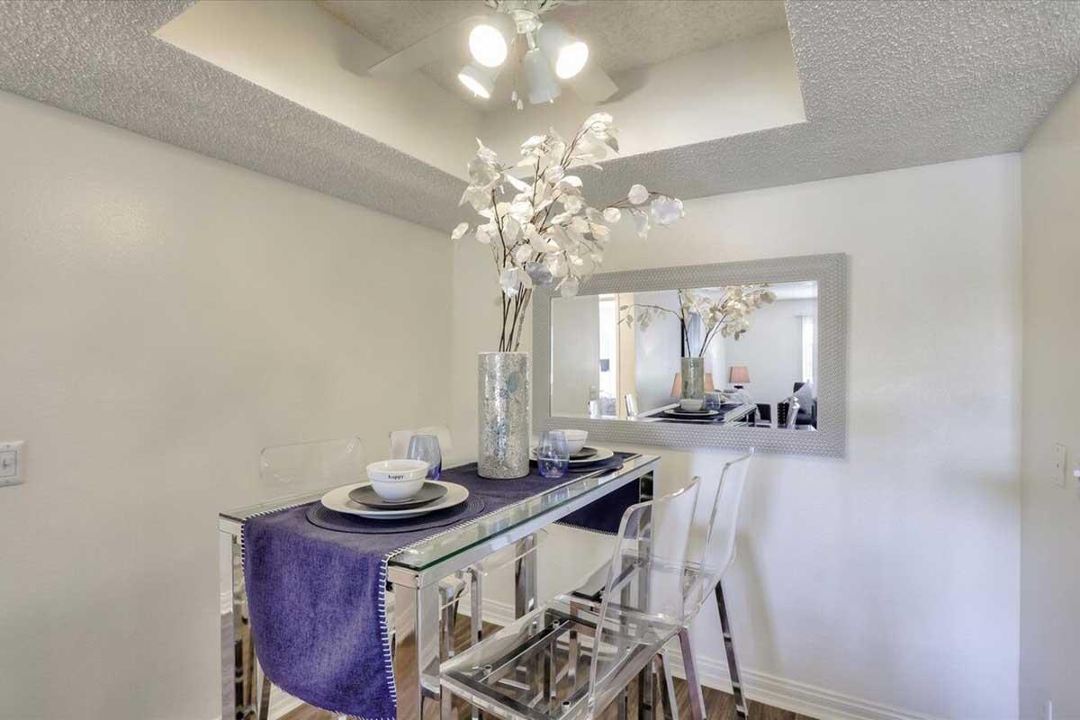 A modern dining area featuring a small glass table with clear chairs, adorned with a purple table runner. A decorative vase with white flowers sits on the table, and a mirror reflects the room's light. The walls are painted white, and the ceiling has recessed lighting.