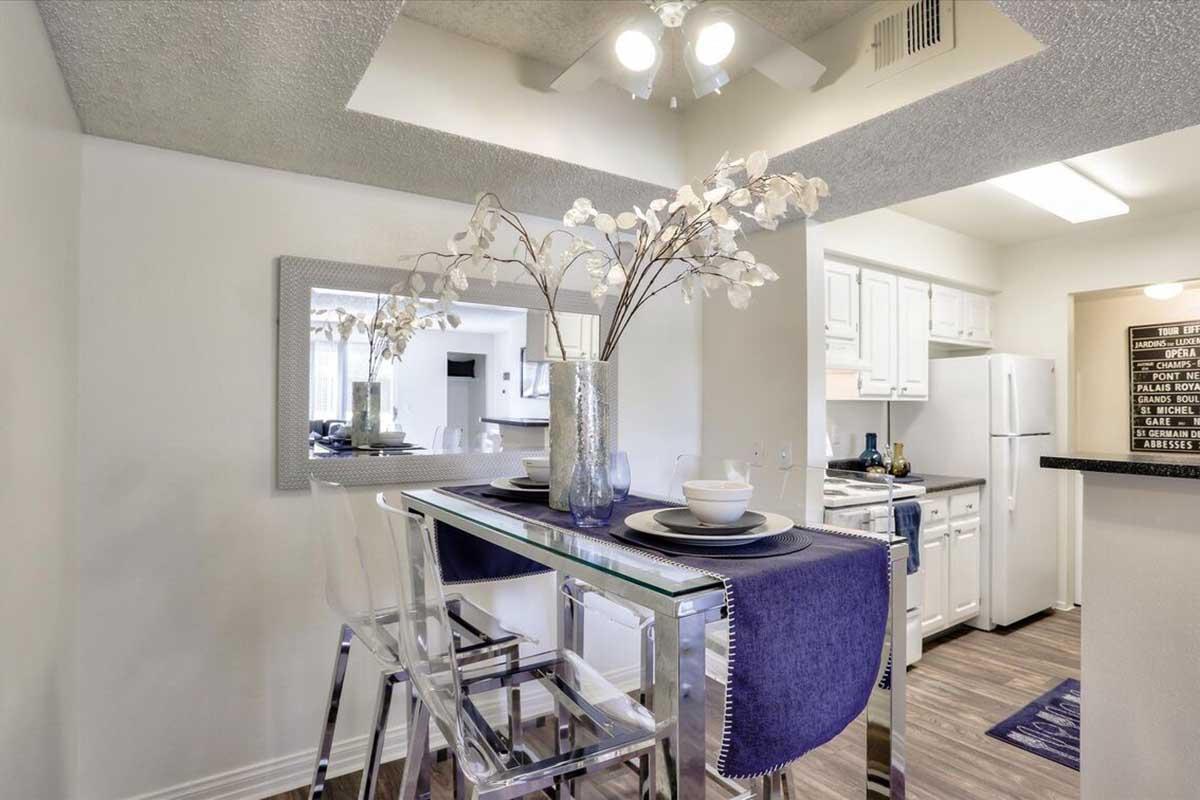 A modern kitchen space featuring a glass-top dining table with clear chairs, adorned with a vase of decorative branches. The kitchen has white cabinets, a dark countertop, and stainless steel appliances, with a mirror reflecting the room's light. A soft purple runner adds a pop of color.