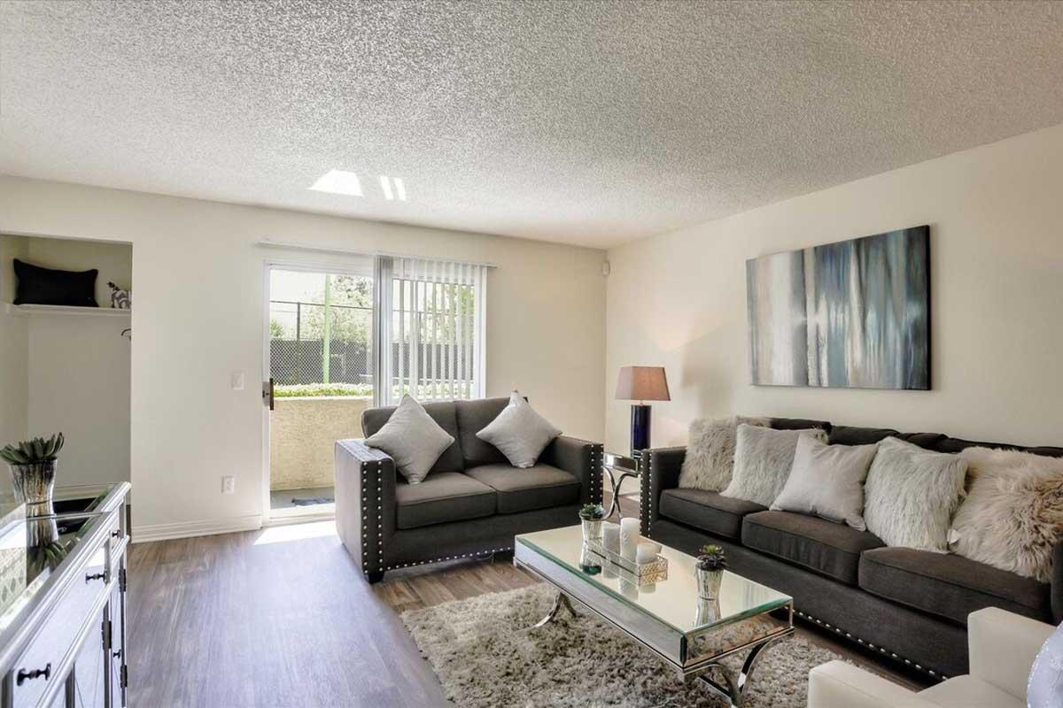 A cozy living room featuring a light-colored wall, two gray sofas with decorative pillows, a glass coffee table, and a lamp. Natural light streams in through sliding glass doors that lead to a small outdoor space. Decorative touches include a piece of abstract art and a potted plant on a side table.