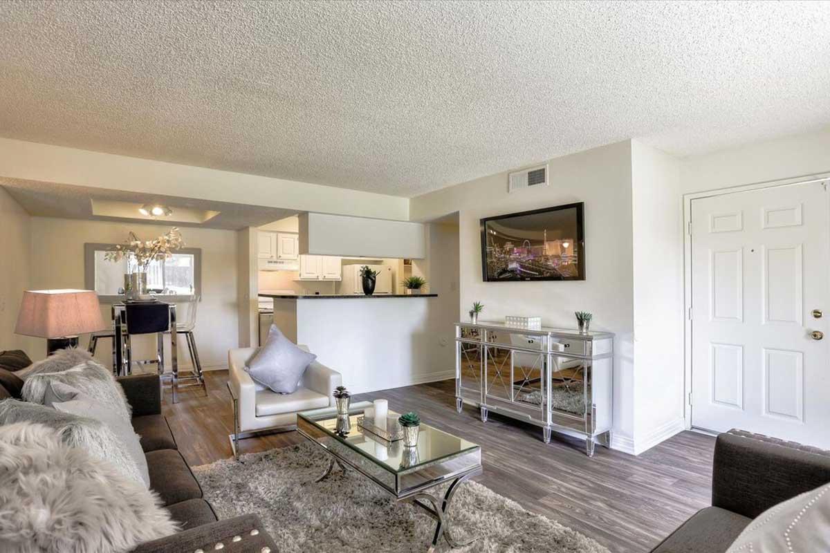 A bright living room featuring a comfortable gray sofa with plush pillows, a glass coffee table, and a stylish silver console table. There is a small dining area with bar stools and a white kitchen visible in the background. The walls are light-colored, and the space is well-lit with natural light.