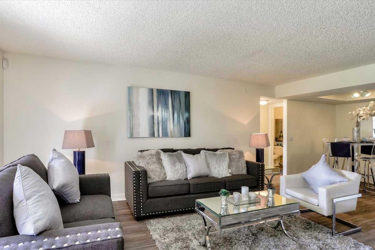 A cozy living room featuring a large gray sofa with decorative pillows, a white accent chair, and a glass coffee table. Two stylish lamps provide soft lighting, and a colorful abstract painting adorns the wall. The space has a warm, inviting atmosphere with a carpeted floor and an open layout leading to a dining area.