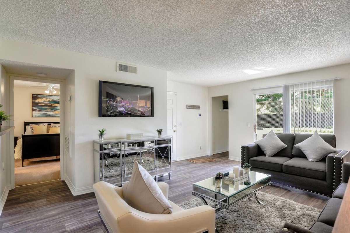 A modern living room featuring a grey sofa with decorative pillows, an armchair, and a glass coffee table set on a wood-style floor. There's a large wall-mounted TV and a mirrored console table. Natural light streams in through a window, enhancing the airy feel of the space.
