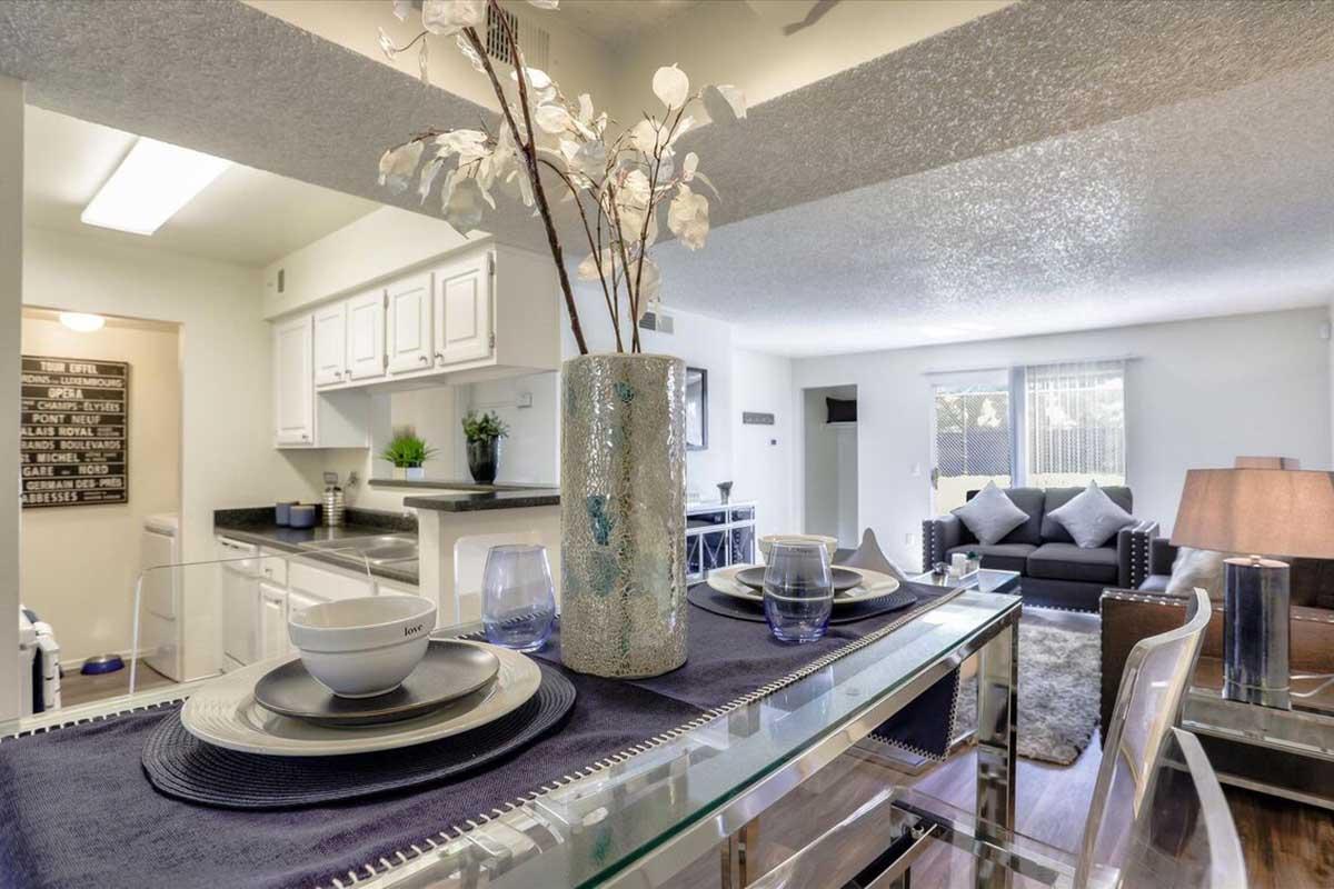 A modern dining area featuring a table set with elegant plates and glasses, a decorative vase with white flowers, and a stylish lamp. In the background, a cozy living room with a gray sofa and large windows, along with a well-lit kitchen area visible. The overall ambiance is contemporary and inviting.