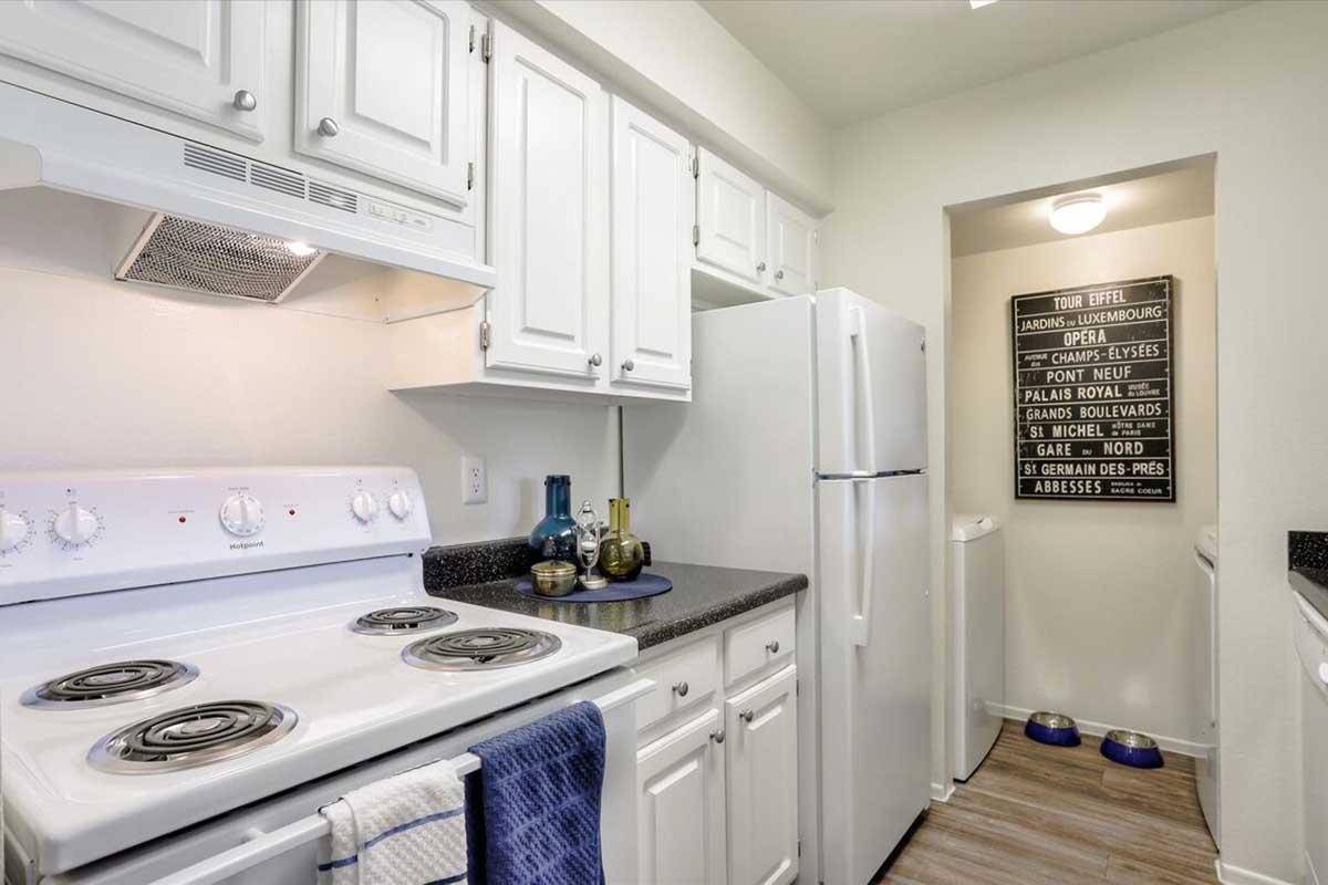 A modern kitchen featuring white cabinets, black countertops, a stove with four burners, a refrigerator, and a small hallway leading to a utility area. Decorative items are on the counter, and a wall sign with various street names is displayed. The flooring is light wood.