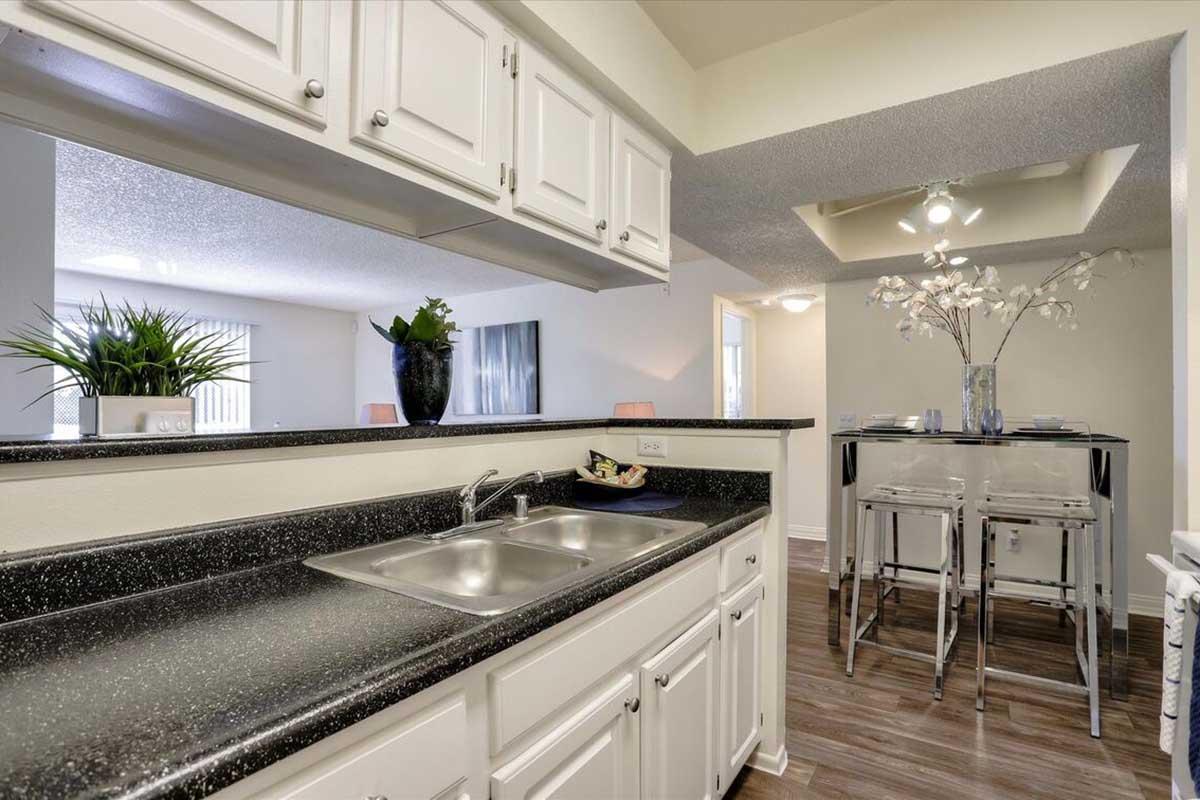 Modern kitchen view with white cabinets and a black countertop, featuring a double sink and decorative plants. A small dining area with clear stools is visible in the background, along with a bright, airy living space. Soft lighting creates a welcoming atmosphere.