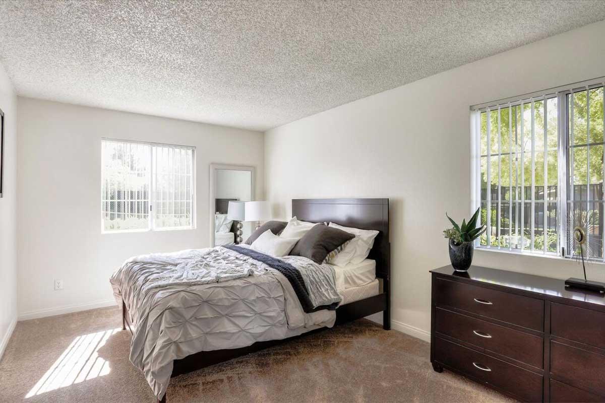 A modern bedroom featuring a bed with grey and white bedding, a nightstand with a lamp, and a dresser. There are two windows allowing natural light, and greenery visible outside. The walls are painted white, and the carpet is light-colored, giving the room a bright and inviting atmosphere.