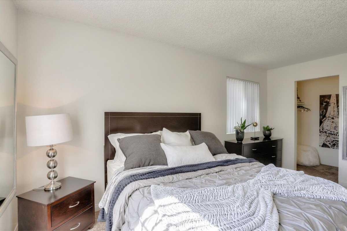 A cozy bedroom featuring a dark wood bed with white and gray bedding. There’s a bedside table with a lamp, and potted plants on top. A dresser is visible on the opposite side, and a closet is seen in the corner. Natural light illuminates the space, creating a warm and inviting atmosphere.