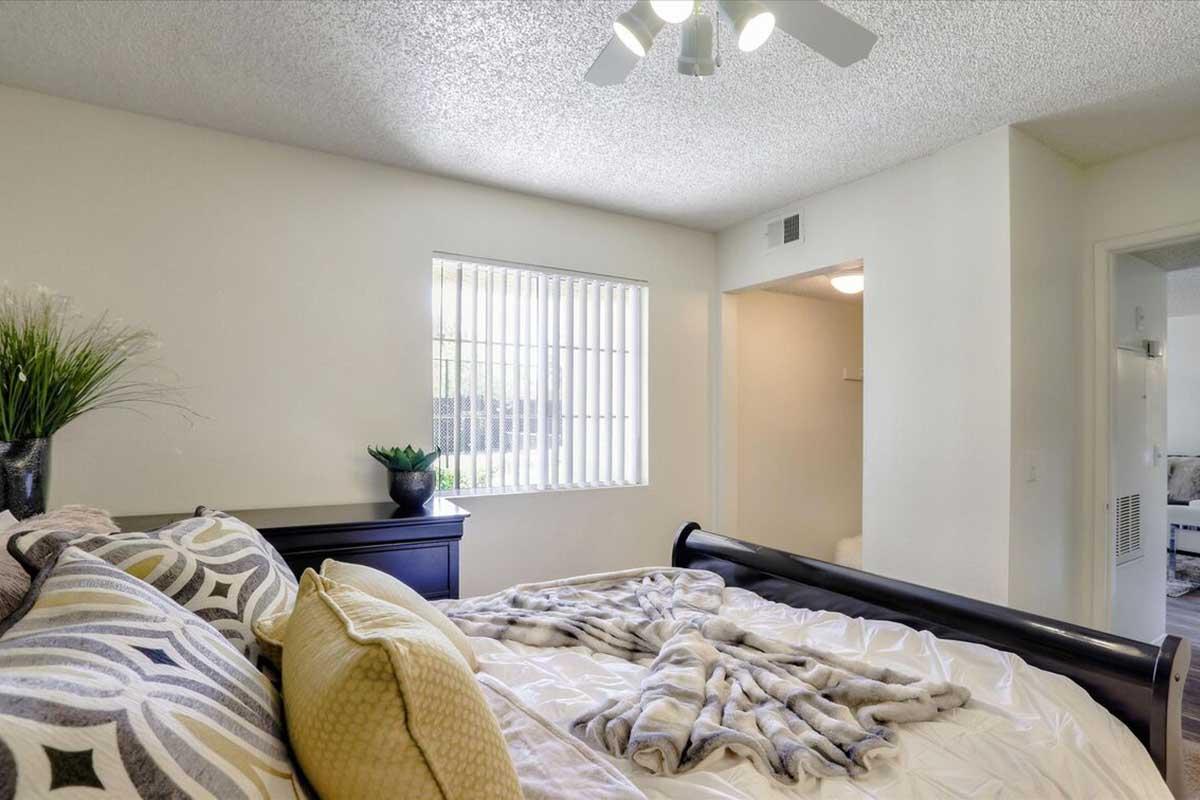 A cozy bedroom featuring a bed with decorative pillows and a soft throw blanket. The room has a window with vertical blinds, allowing natural light to enter. A small potted plant decorates the bedside table, and a ceiling fan is visible above, adding to the inviting atmosphere.