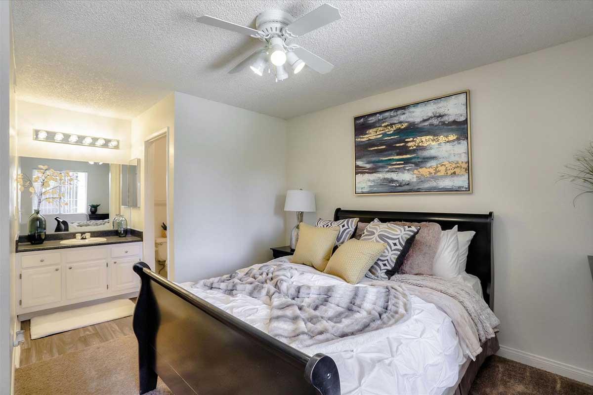A cozy bedroom featuring a dark wooden sleigh bed with white and gray bedding, adorned with yellow accent pillows. A decorative painting hangs above the bed. To one side, there is a vanity area with two sinks, stylish lighting, and a plush rug on the floor, creating a warm and inviting atmosphere.
