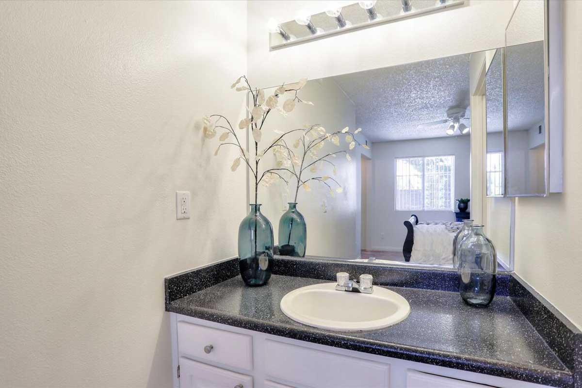 A small bathroom with a dark countertop, featuring a round sink and a faucet. Decorative glass vases with faux white branches are on the countertop. A mirror is mounted above the sink, and in the background, a living space with a bed and a window is visible. The walls are painted a light color.