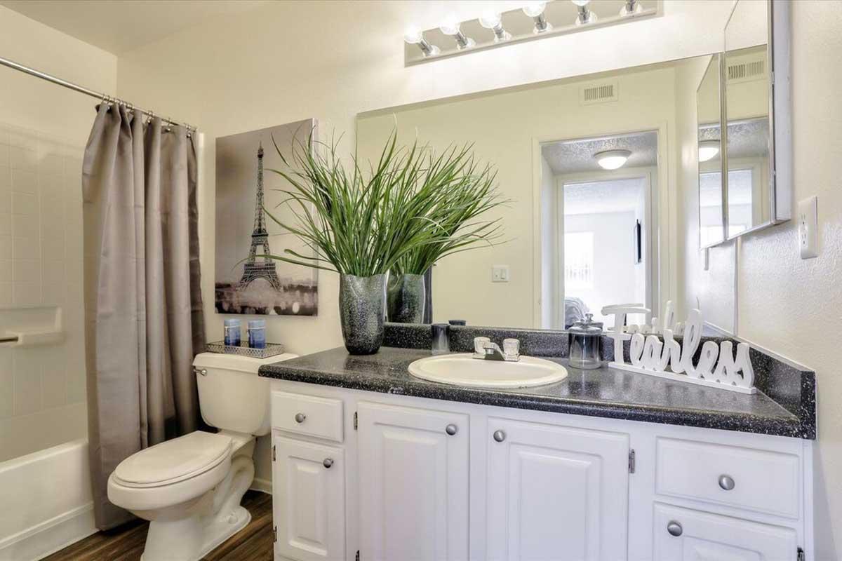A modern bathroom featuring a beige and gray color scheme. The vanity consists of white cabinets with a dark countertop. On the countertop, there is a decorative plant in a silver vase, a decorative sign that reads "Fearless," and toiletries. A shower with a glass door and a photo of the Eiffel Tower is visible on the wall.