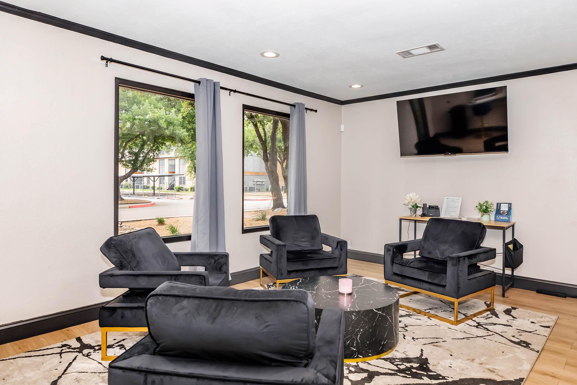 A modern living area featuring two large windows with gray curtains, a black marble coffee table, and four plush black armchairs. There's a TV mounted on the wall and a small table with documents. The space is well-lit, creating an inviting atmosphere.