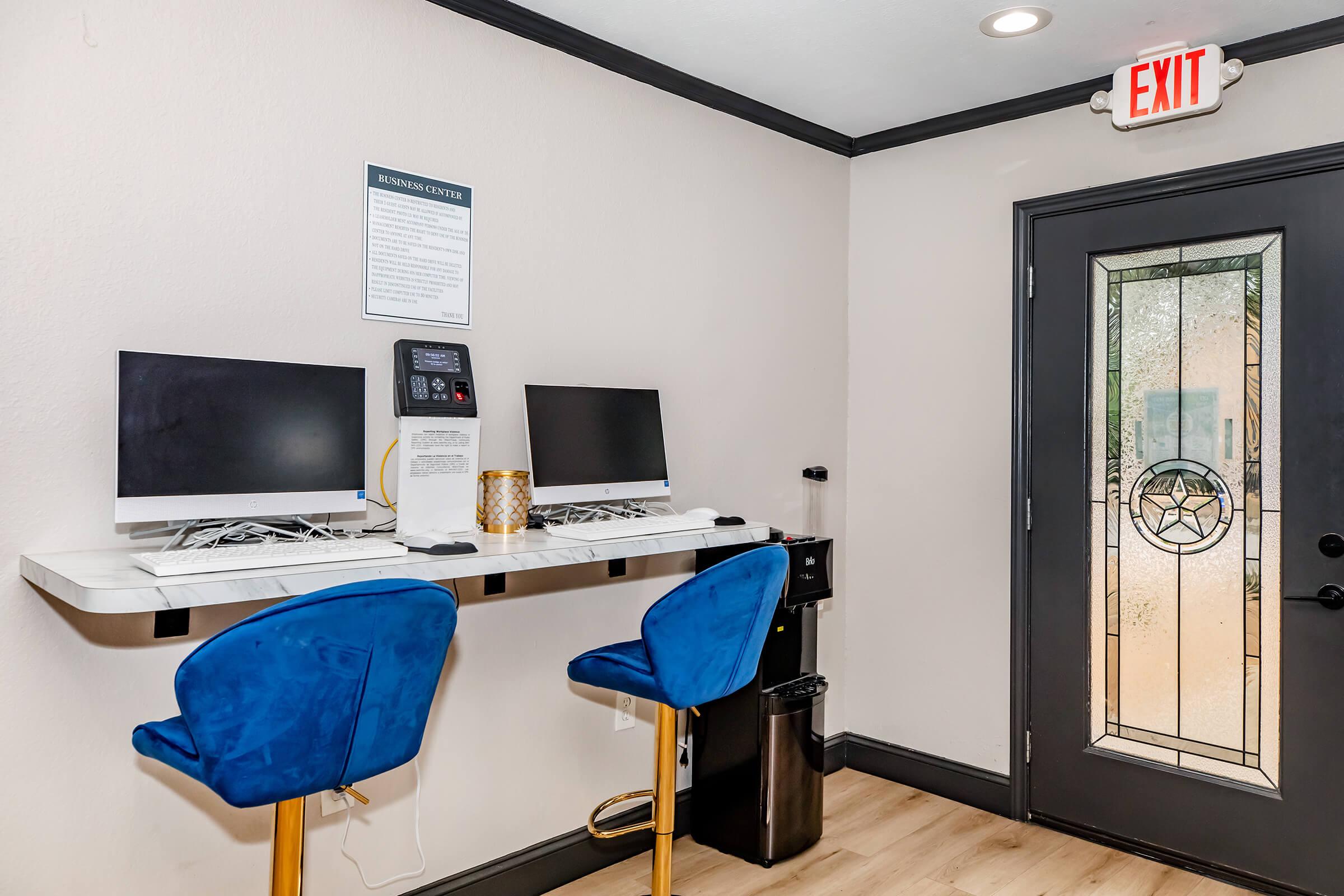 A small workspace featuring two computers on a countertop, with blue stools. There's a decorative gold candle holder, a printer, and a door with a frosted glass panel. A sign on the wall provides information about the center, and an exit sign is visible above the door. The flooring is light wood.