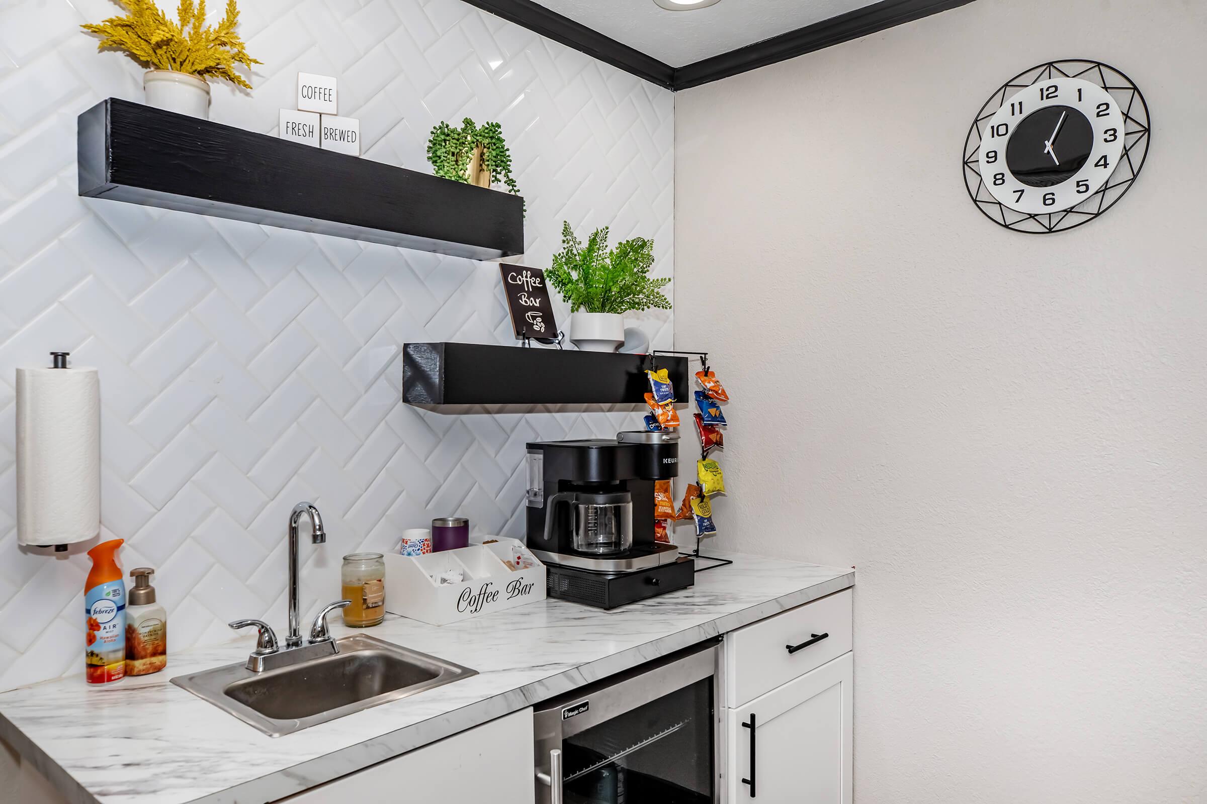 A cozy coffee corner featuring a black coffee maker on a marble countertop, surrounded by various coffee supplies and snacks. A small sink is visible, along with a paper towel dispenser and decorative plants. The wall has a stylish clock and a herringbone-patterned backsplash, adding warmth to the space.