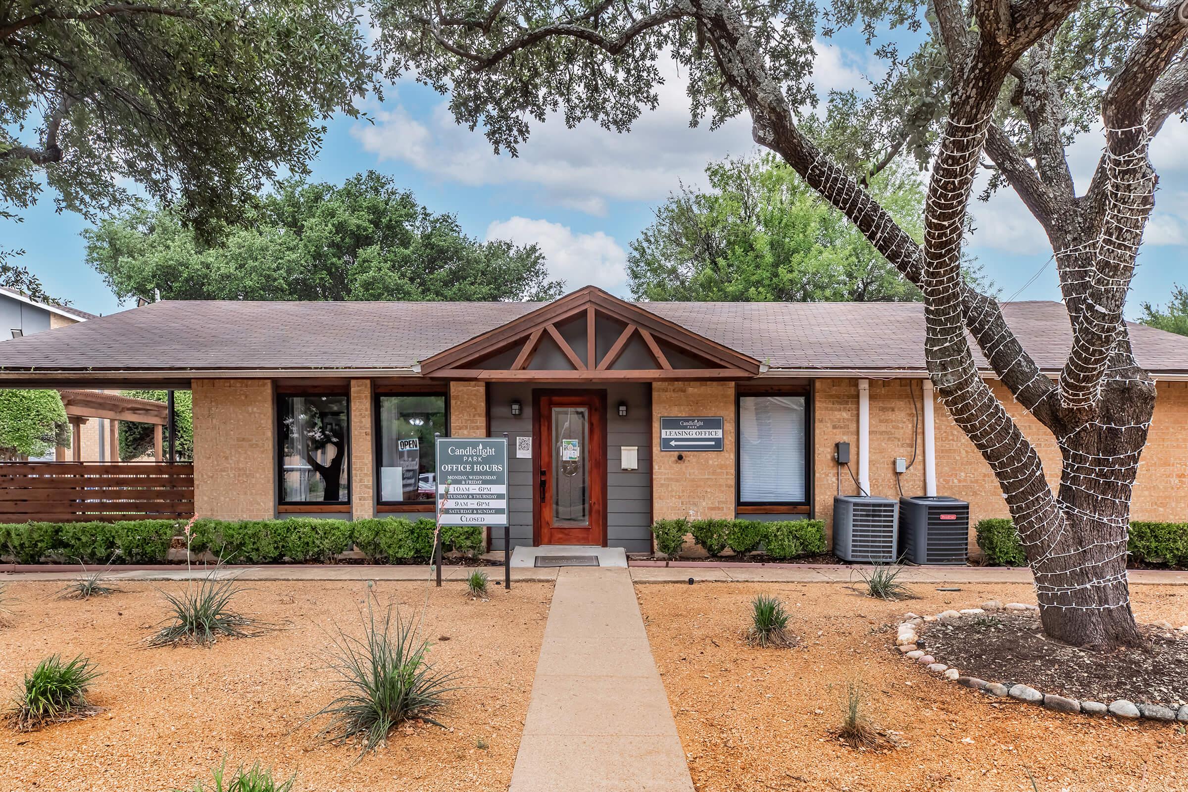 A charming single-story building with a brown and gray exterior, featuring a peaked roof and a welcoming entrance. Flanked by greenery and landscaping, the front yard includes small bushes and a pathway leading to the door. A large tree with lights is on the right side, enhancing the inviting atmosphere.