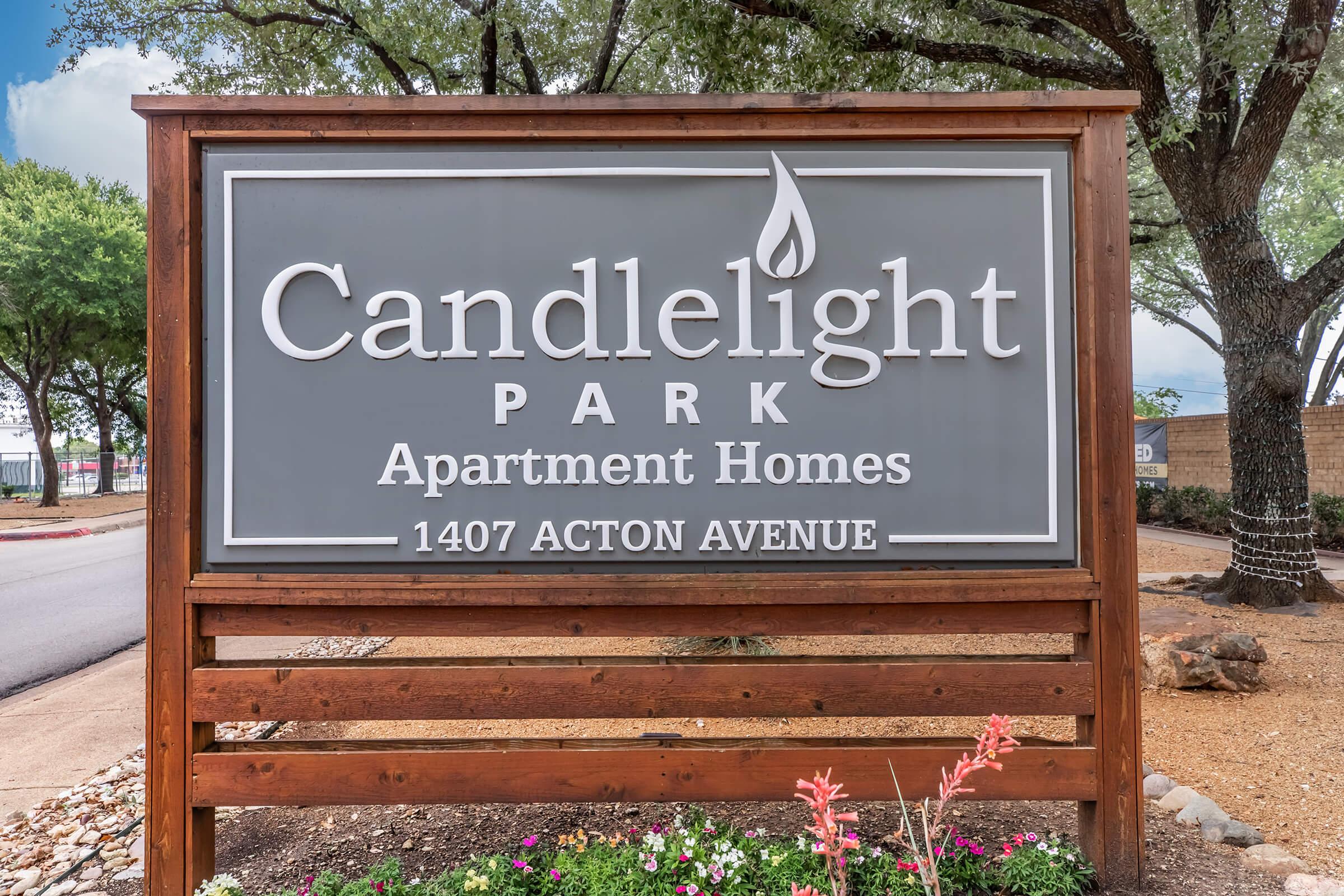 Sign for Candlelight Park Apartment Homes, displaying the name prominently with a logo and the address 1407 Acton Avenue. Surrounded by greenery, including trees and flowering plants in the foreground, set against a clear sky.