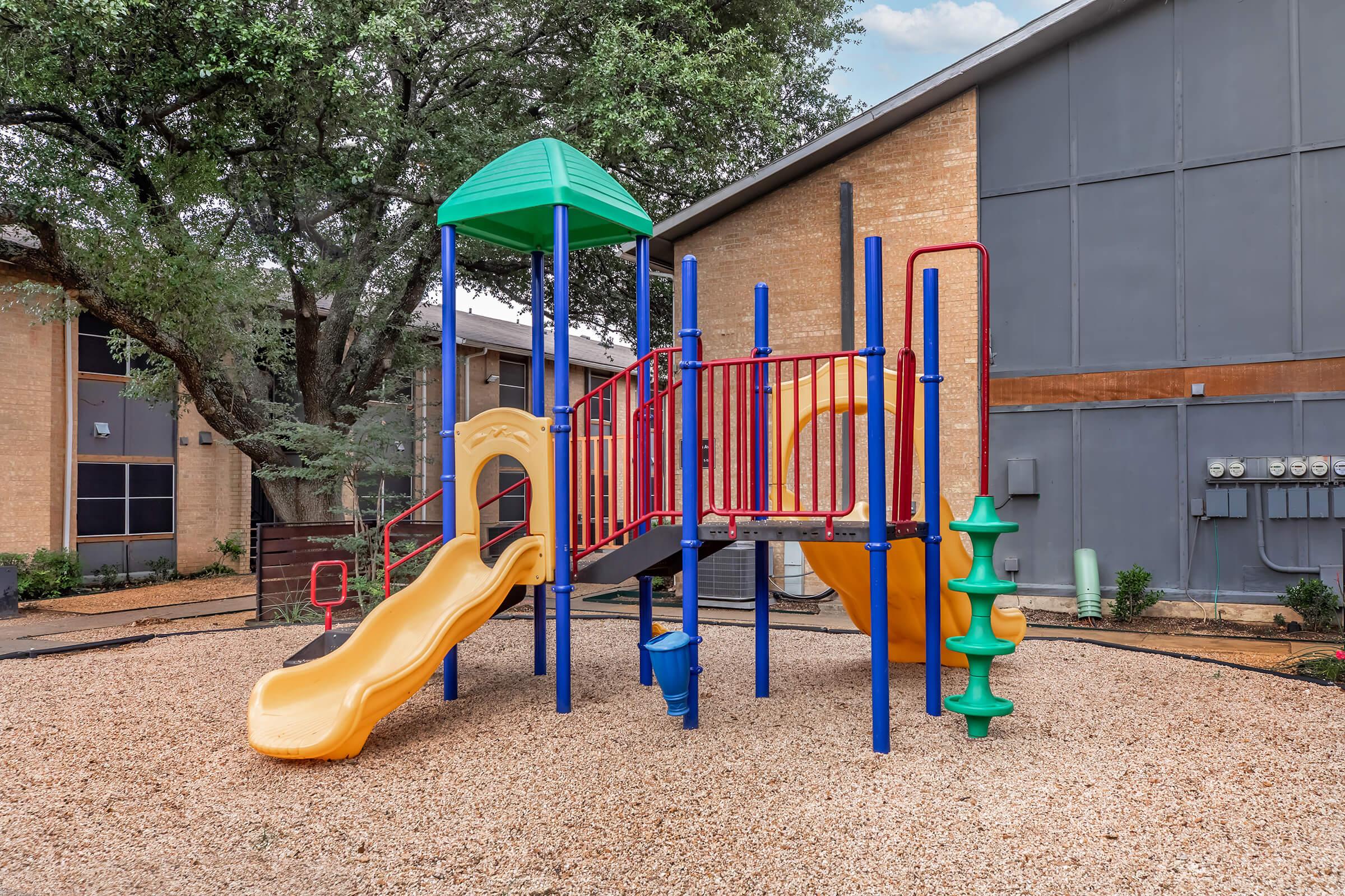 A colorful playground featuring a yellow slide, climbing structure, and a green roof. The playground is surrounded by gravel and is located next to a brick building with a modern design. Trees are visible in the background, providing shade.