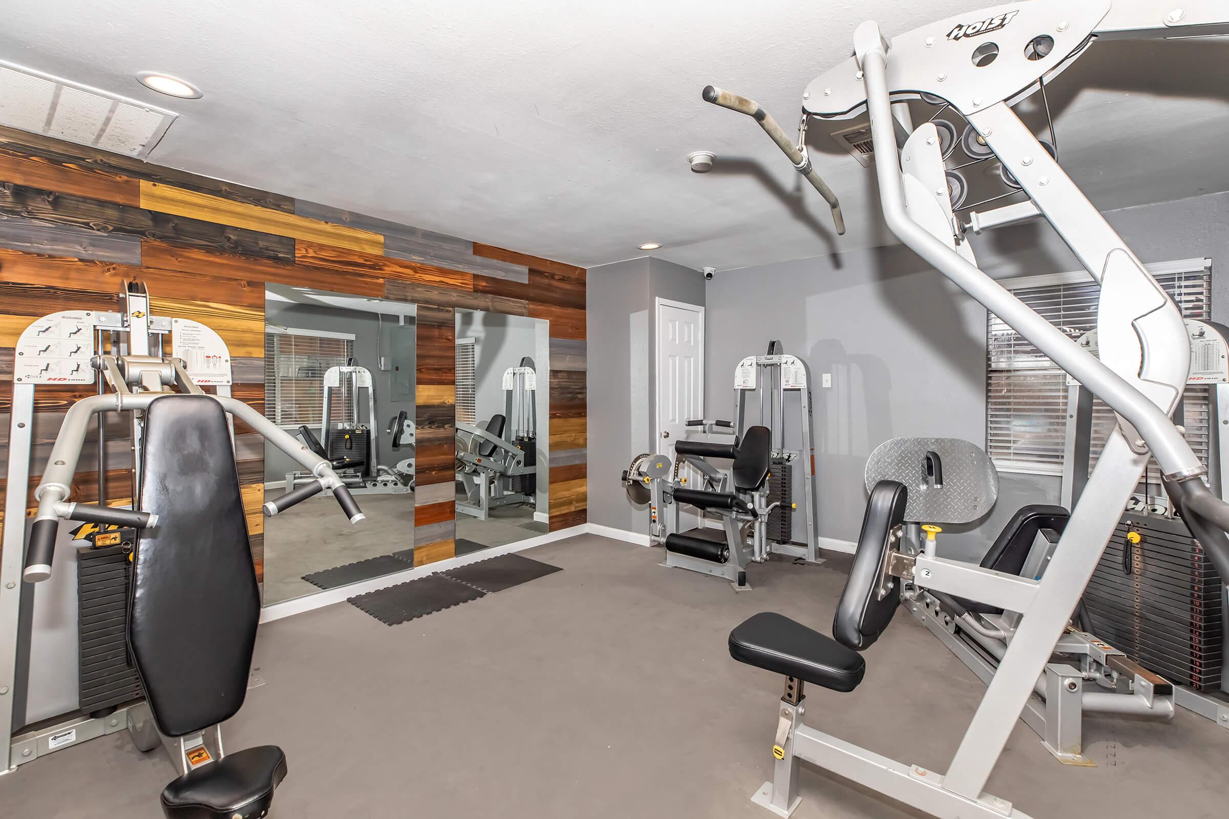 A modern home gym featuring various exercise equipment, including weight machines and benches, set against a wall of wooden panels. A large mirror reflects the gym space, enhancing the sense of openness. The flooring is carpeted, creating a comfortable workout environment.