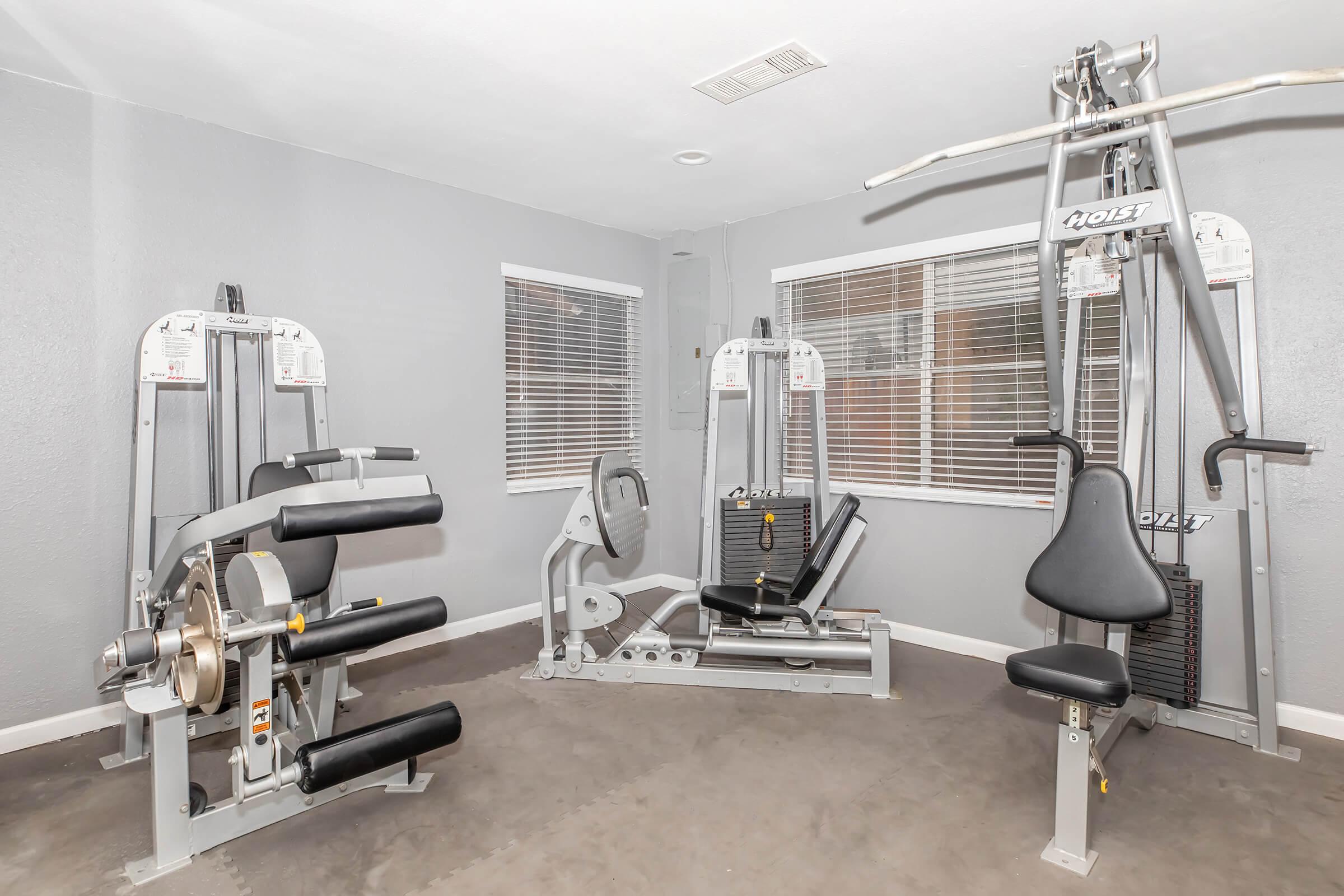 A modern home gym featuring multiple exercise machines, including a cable pulley system, a leg press, and a rowing machine, set against gray walls with windows lined with blinds. The floor is carpeted, providing a comfortable workout environment.