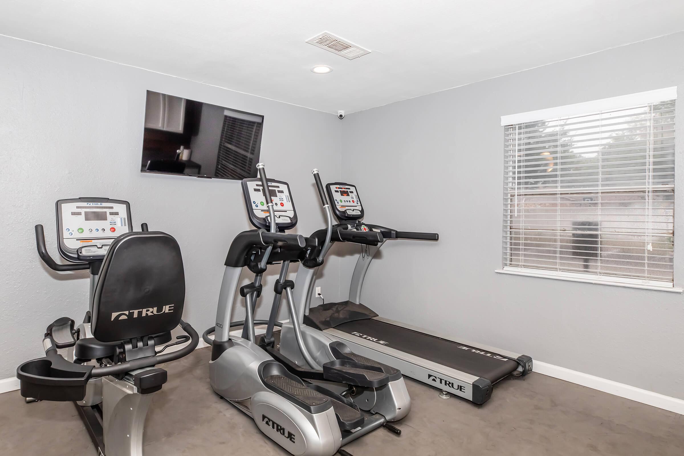 A small fitness room featuring two exercise bikes, a treadmill, and an elliptical machine. There's a wall-mounted television and a window with blinds letting in natural light. The walls are painted in a neutral tone, and the floor is carpeted.