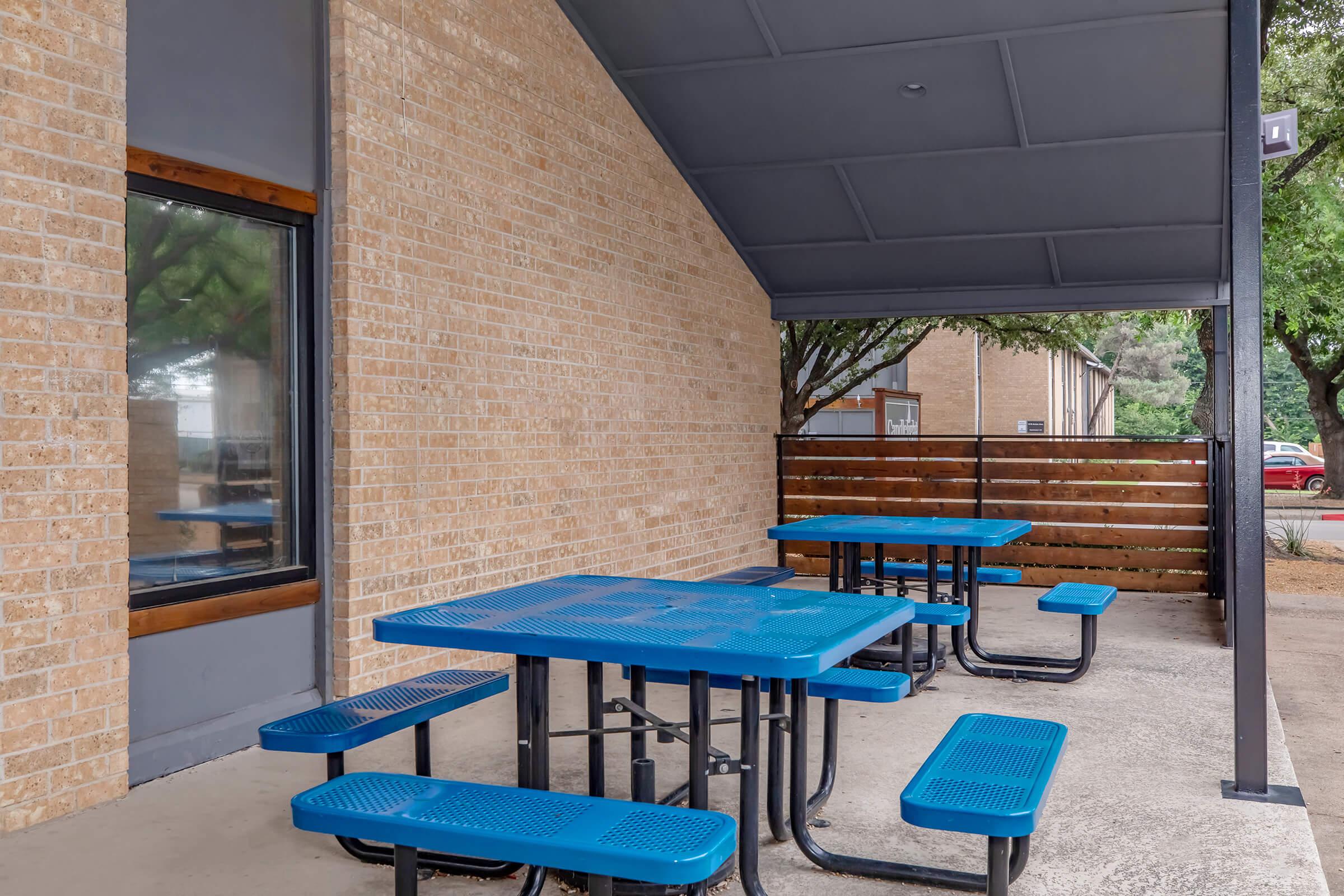 An outdoor seating area featuring a blue picnic table with attached benches, set against a brick wall and a shaded overhang. Trees are visible in the background, providing a natural environment, and a wooden fence lines the area.