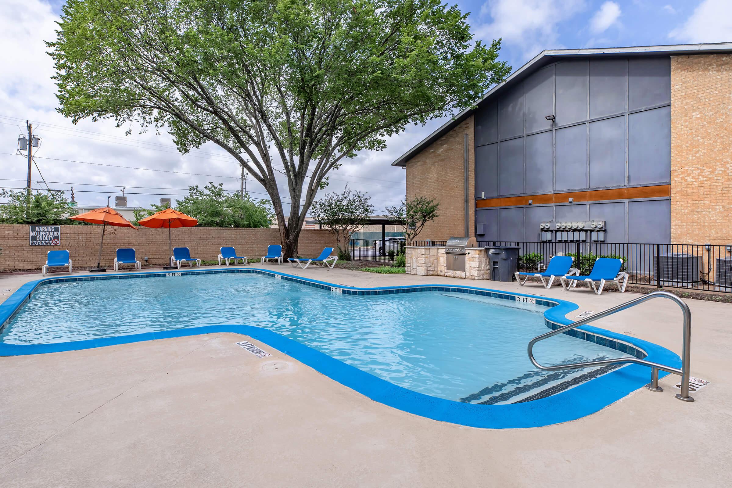 A clean, inviting outdoor swimming pool surrounded by lounge chairs and orange umbrellas. A large tree provides shade nearby, and a brick building with a modern design is in the background. The pool area is well-maintained, creating a relaxing atmosphere for leisure and recreation.