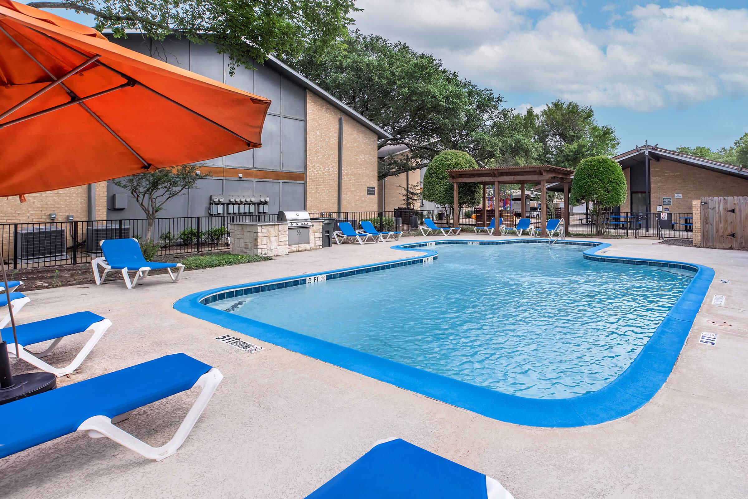 A clear blue swimming pool surrounded by lounge chairs and an orange umbrella. In the background, there are trees, a barbecue area, and a building. The pool area is inviting and well-maintained, ideal for relaxation and outdoor gatherings.