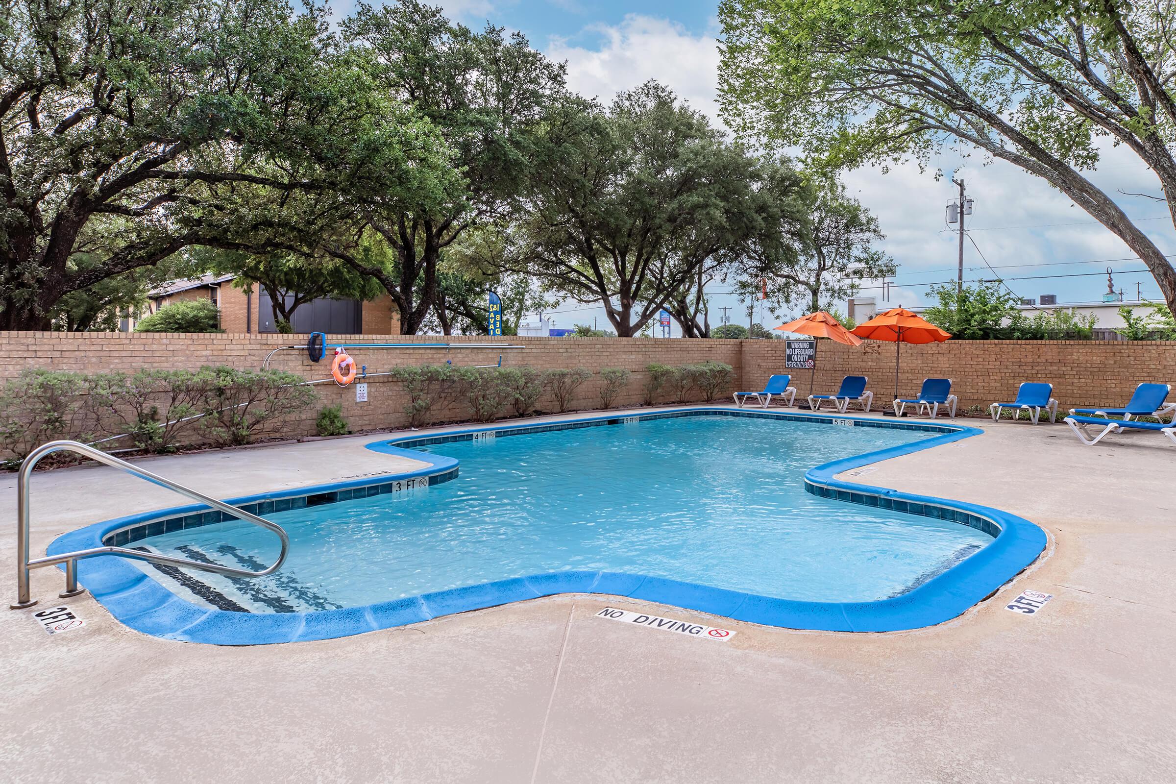 A clean pool surrounded by a patio area featuring lounge chairs, orange umbrellas, and lush greenery. The pool has a unique shape, with clear water and a "No Diving" sign visible. Trees provide shade, and there are safety features like a lifebuoy. The scene is bright and inviting.
