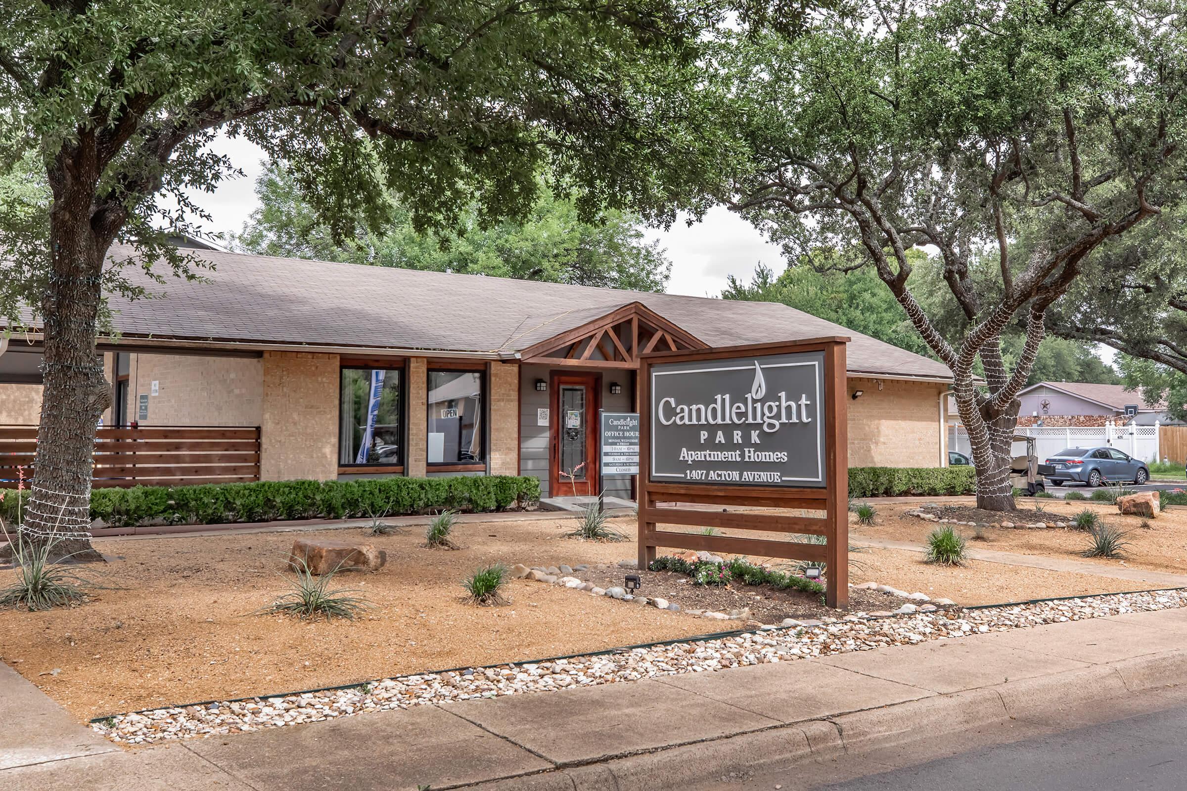 Exterior view of Candlelight Park Apartment Homes, featuring a welcoming sign and landscaping with shrubs and gravel. The building has a sloped roof and is surrounded by trees, providing a pleasant residential atmosphere.