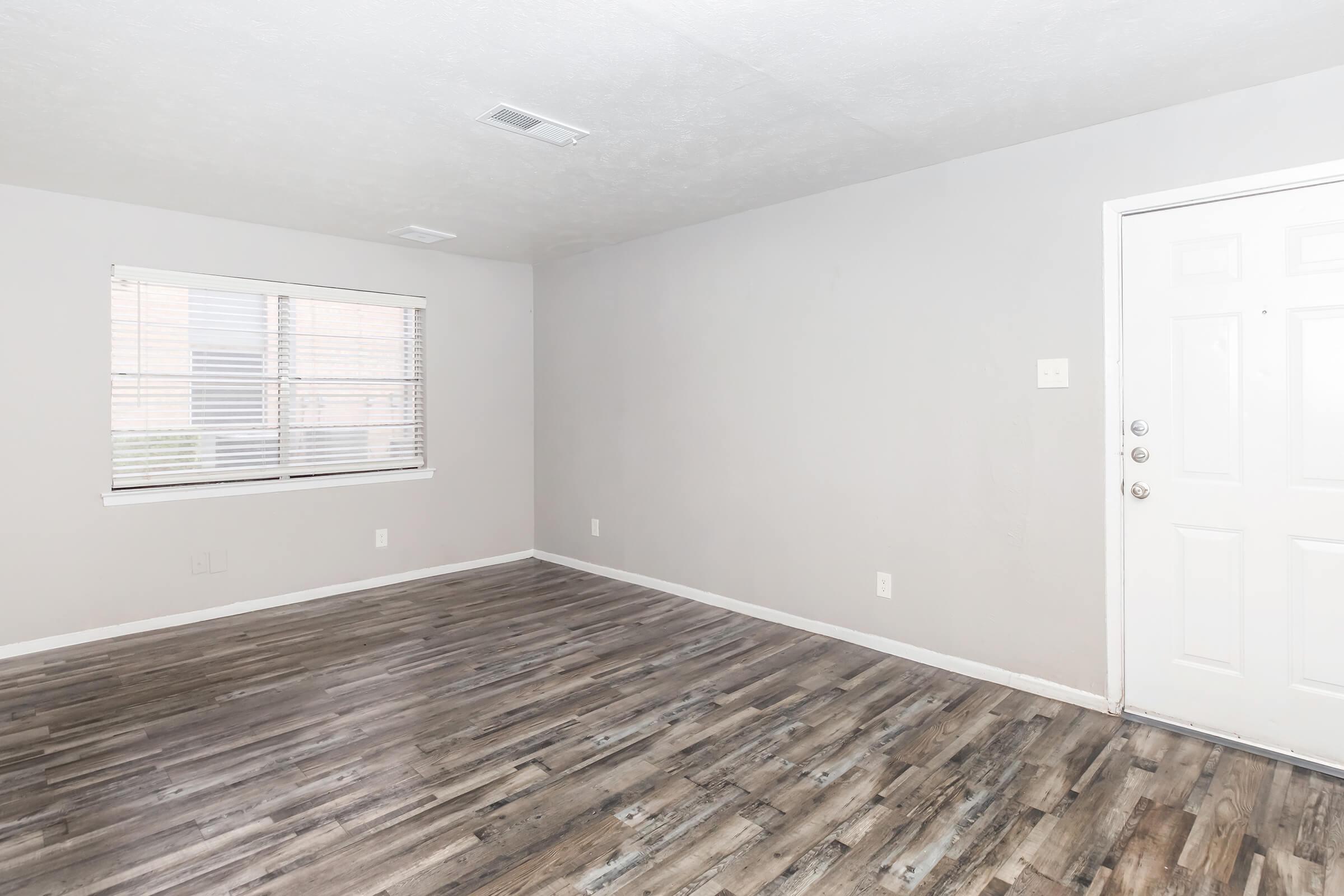 Empty room with light gray walls and laminate flooring. A window with blinds is visible on the left side, allowing natural light into the space. The entrance door is on the right side, featuring a simple design. The overall atmosphere is bright and minimalistic, suitable for various uses.