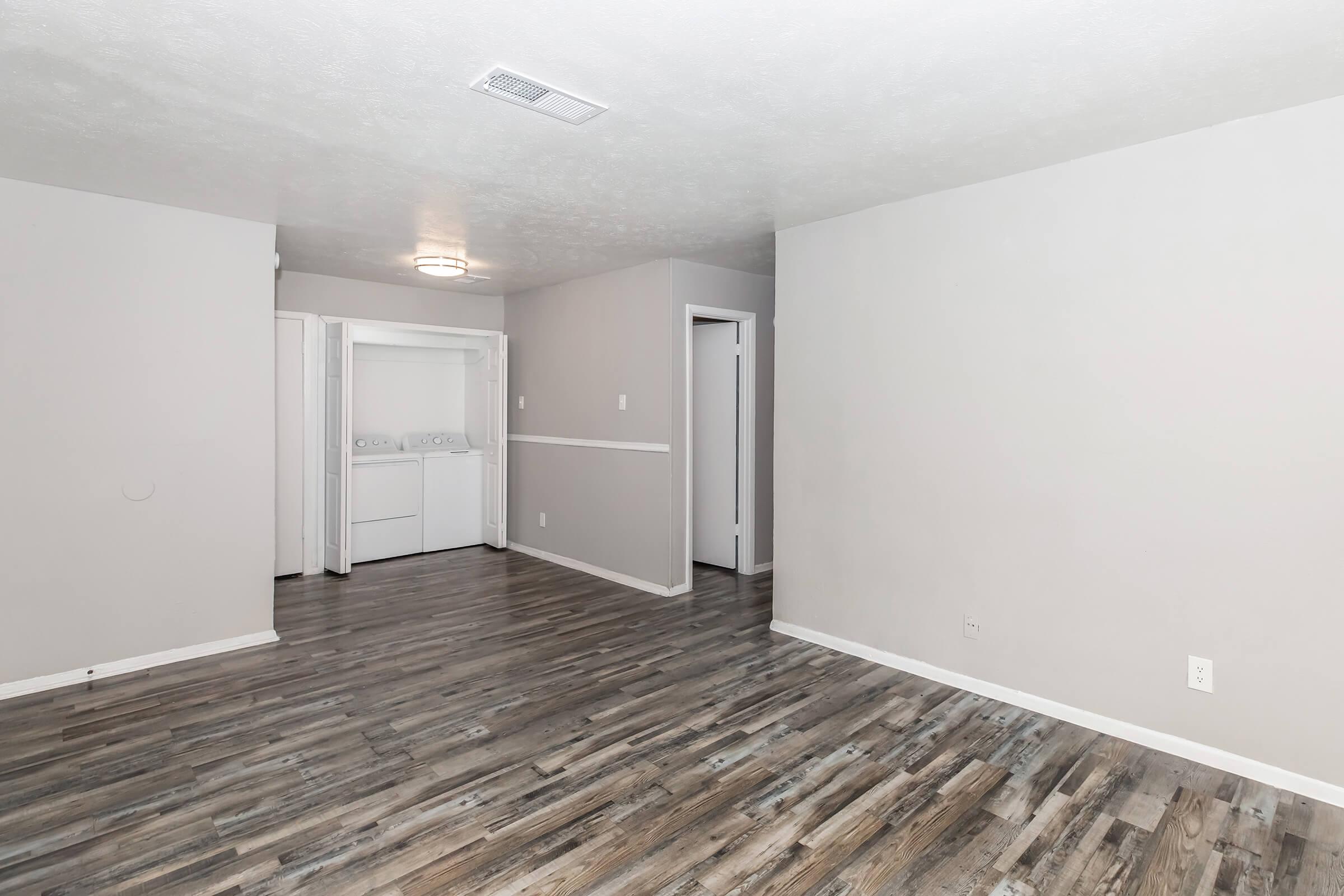 Spacious, empty living area with light gray walls and modern laminate flooring. A laundry closet is visible on the left, and a doorway leads to another room. Natural light fills the space, enhancing the open layout. The ceiling features a simple light fixture, contributing to a bright atmosphere.